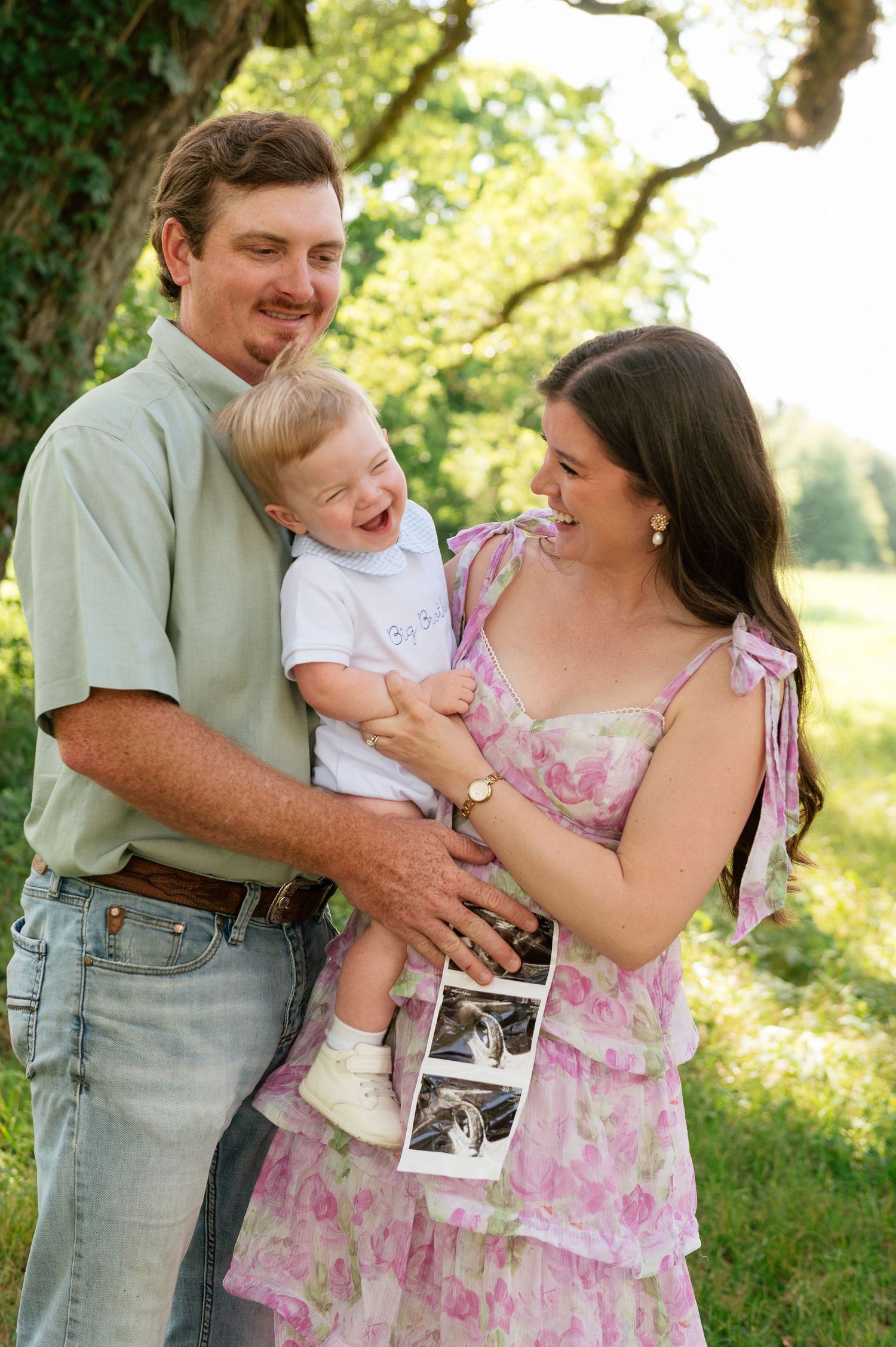 Happy family outdoors, man holding laughing toddler beside pregnant woman in floral dress holding ultrasound photos.