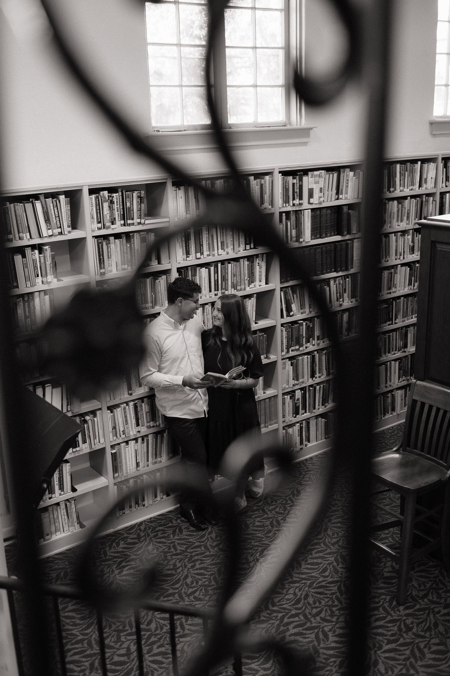 Black and white photo of a couple sharing a book in a classic library surrounded by floor-to-ceiling bookshelves.