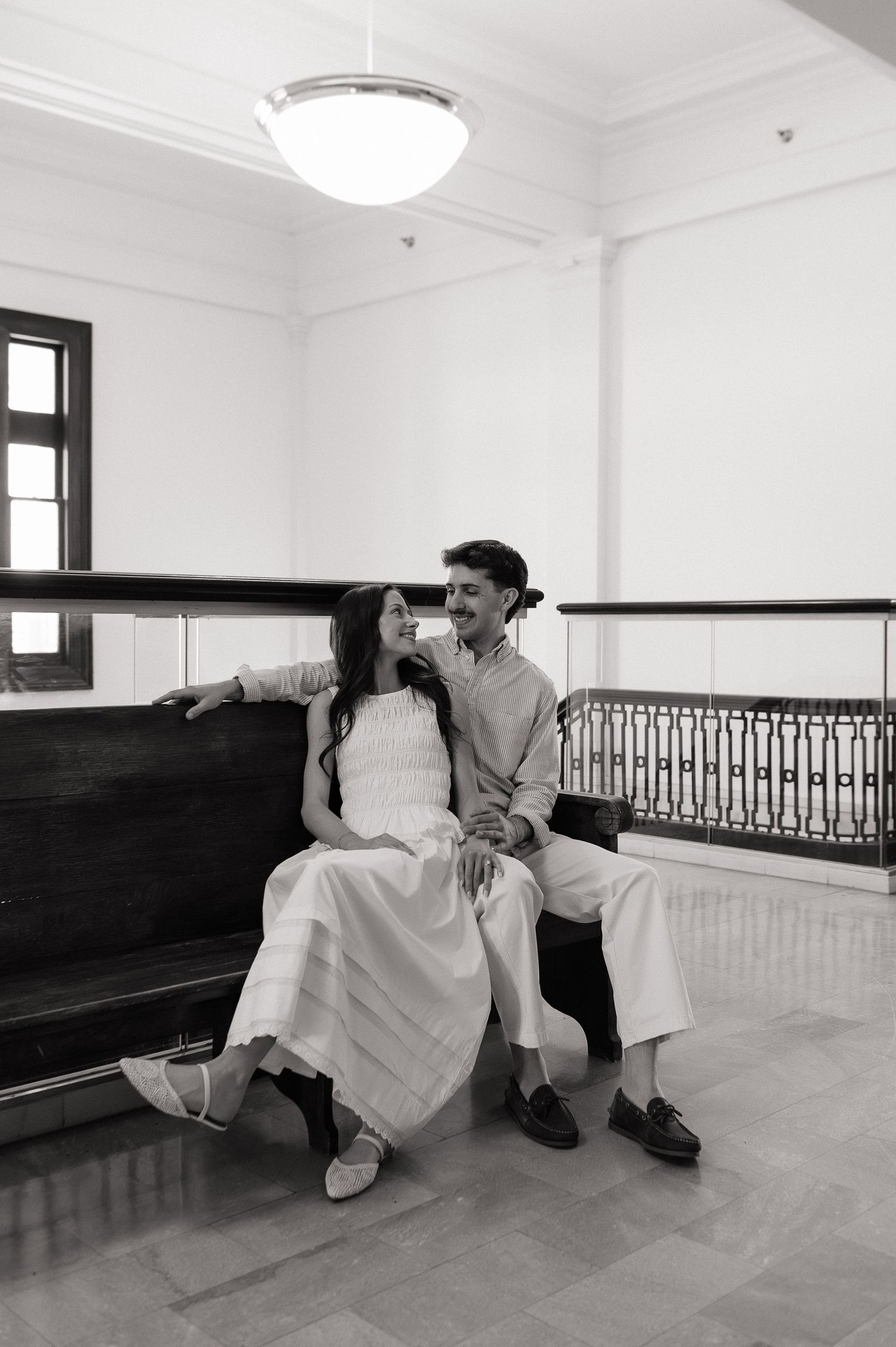 Black and white photo of a couple sitting on a bench in an elegant interior, smiling and holding hands.