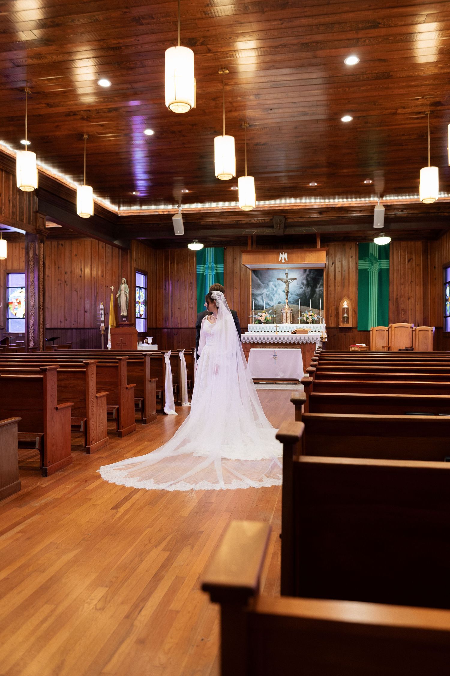 Bride in white gown and long veil walks down wooden church aisle toward altar during wedding ceremony.