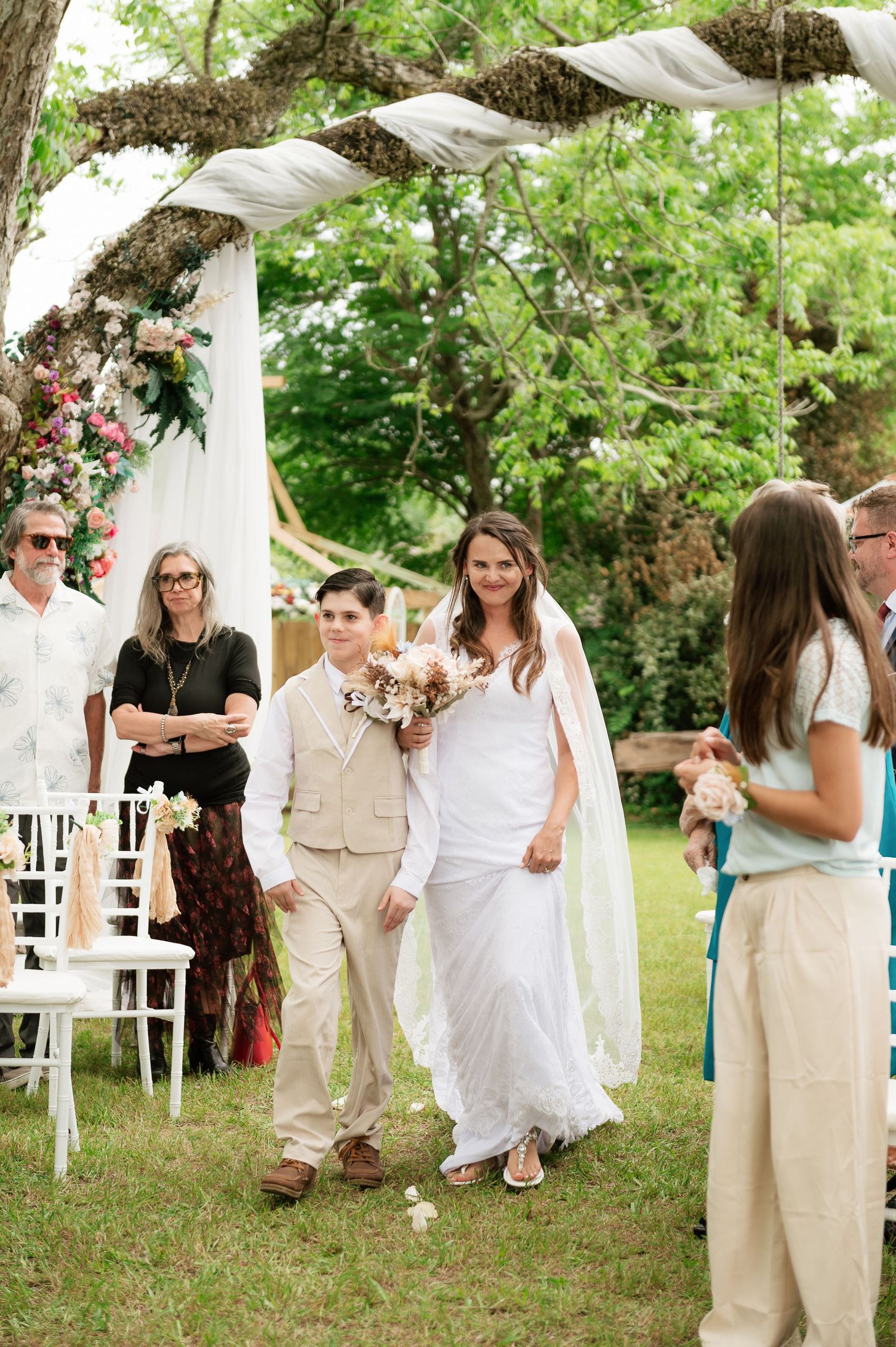 Bride in white gown walks down outdoor aisle with young escort under floral arch at garden wedding.