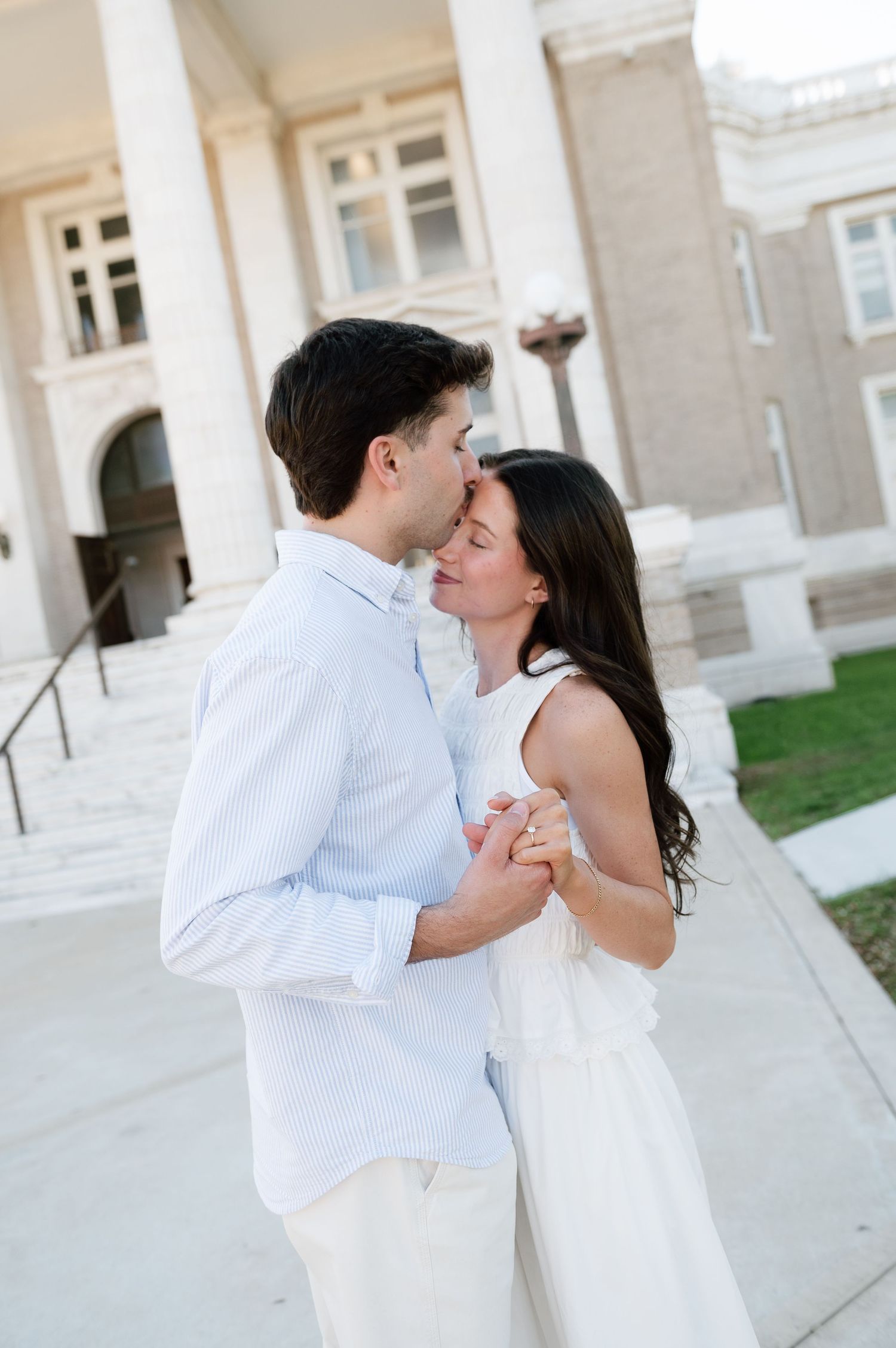 A couple dressed in white shares a romantic kiss while holding hands in front of a classic white building.
