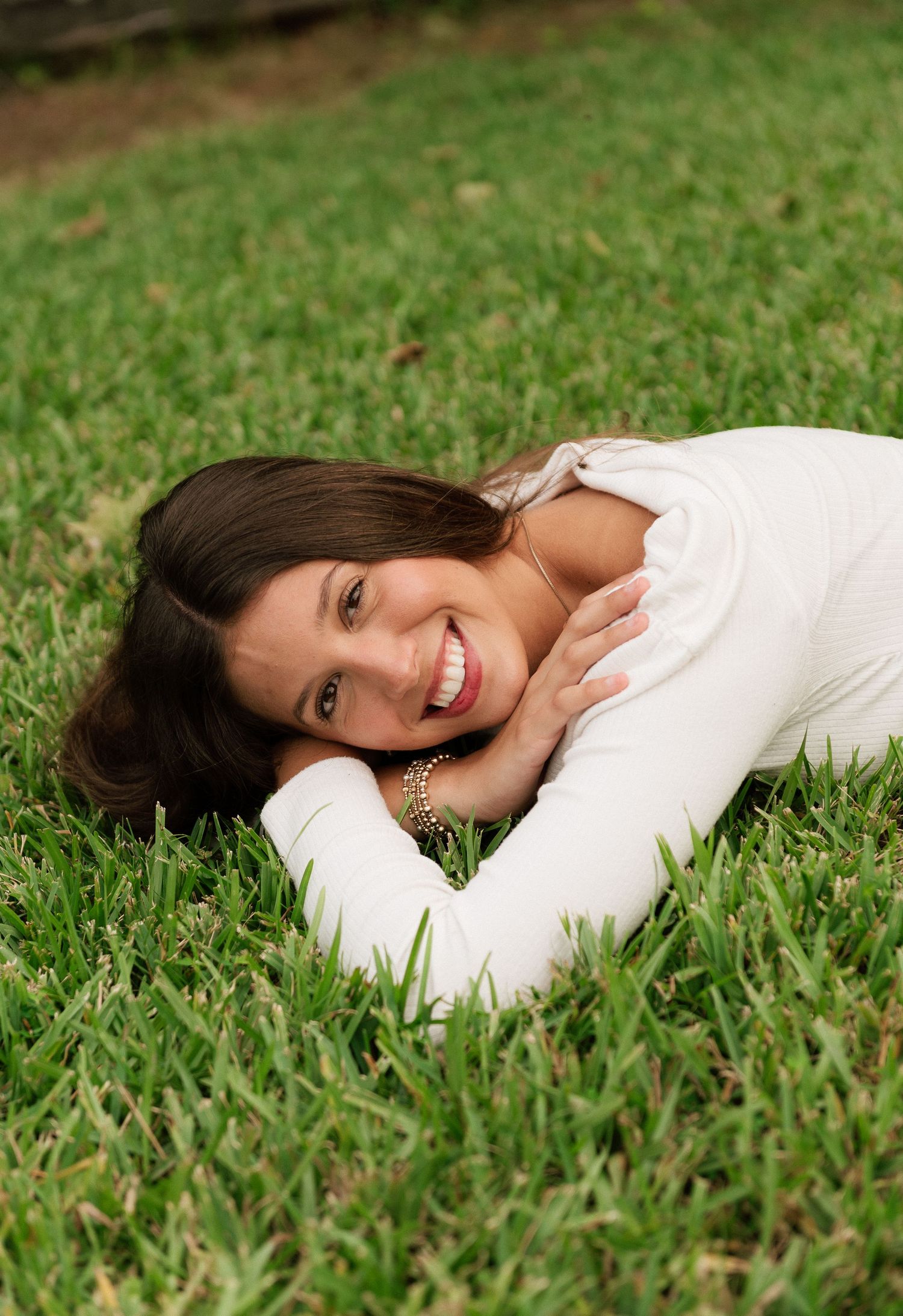 Young woman with dark hair smiling while lying on lush green grass, wearing a white long-sleeve top.