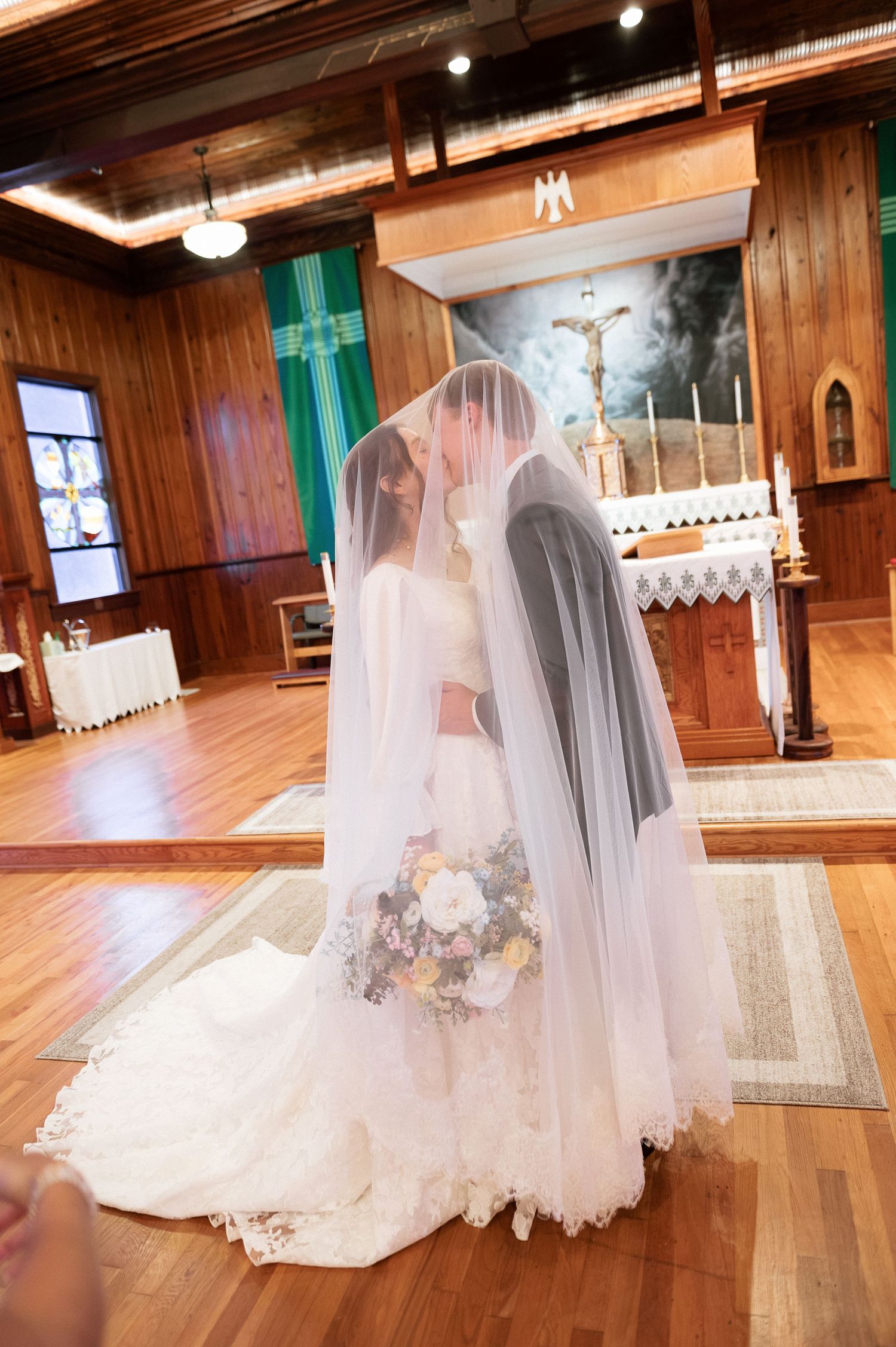 Bride and groom share first kiss under wedding veil at church altar during romantic Catholic wedding ceremony.
