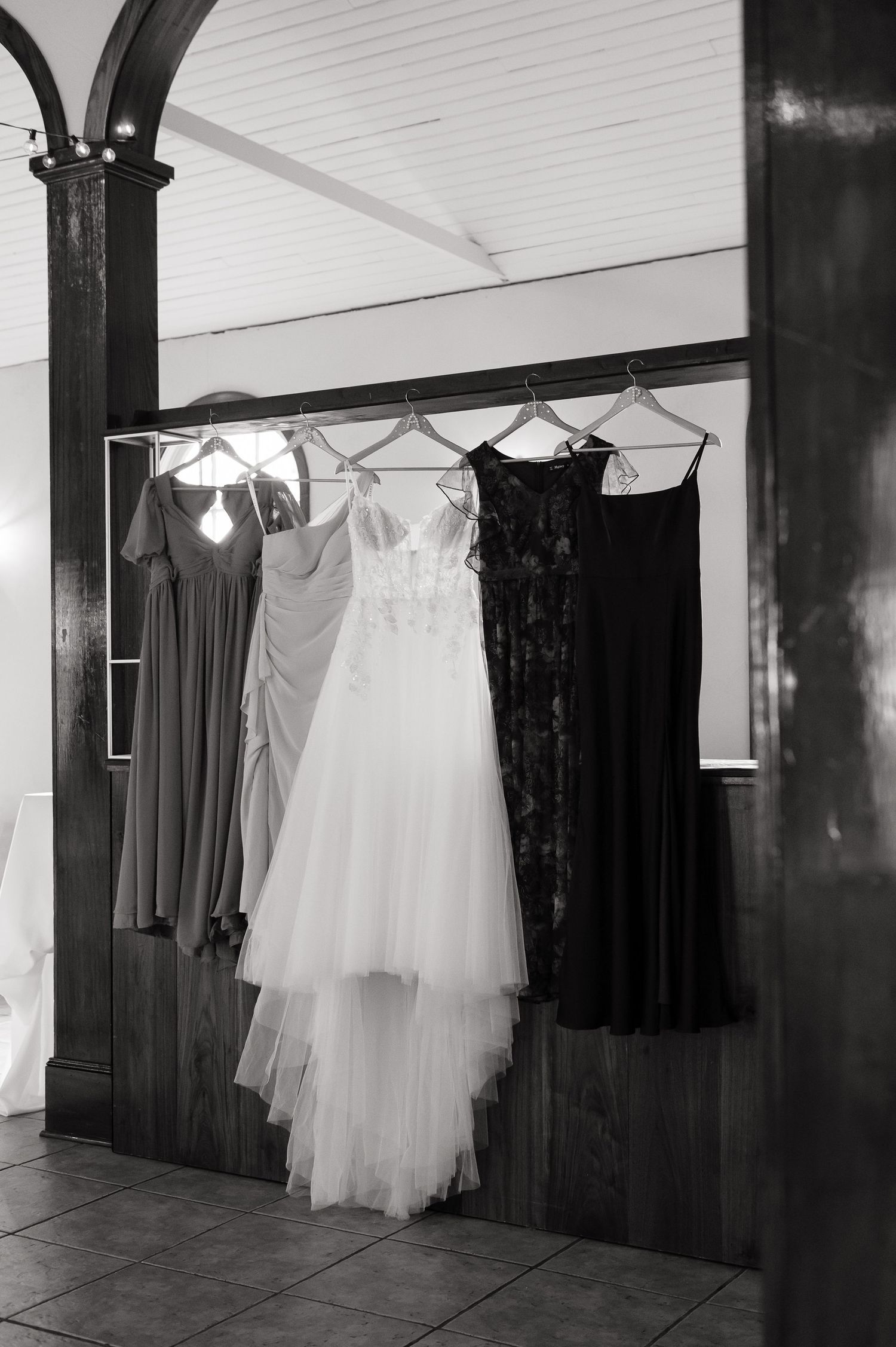 Black and white photo of a wedding dress and bridesmaid gowns hanging on a rack in an elegant venue.