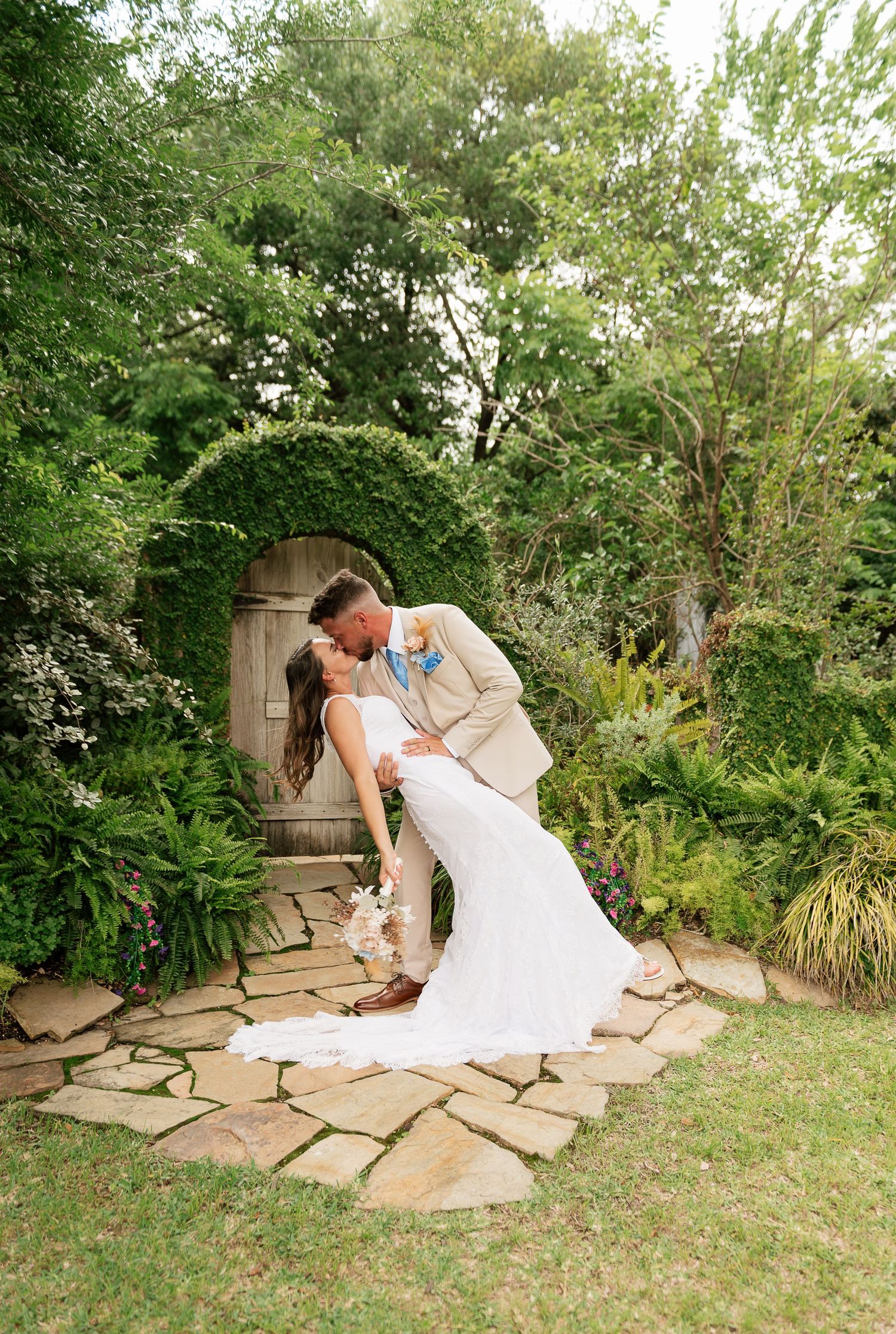 Bride and groom share a romantic dip kiss in a lush garden with an ivy-covered arched wooden door backdrop.