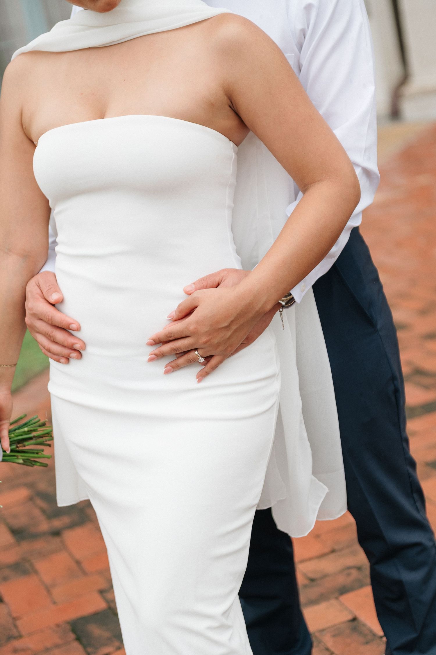 Bride in strapless white sheath wedding dress with groom in navy pants embracing on brick pathway.