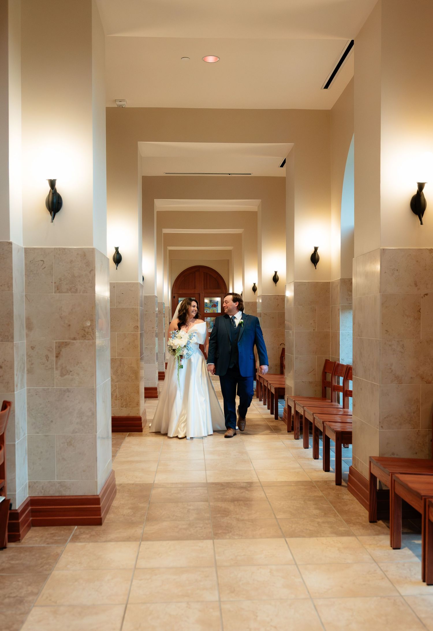 A bride and groom walk hand in hand down an elegant chapel corridor with warm lighting and wooden pews.