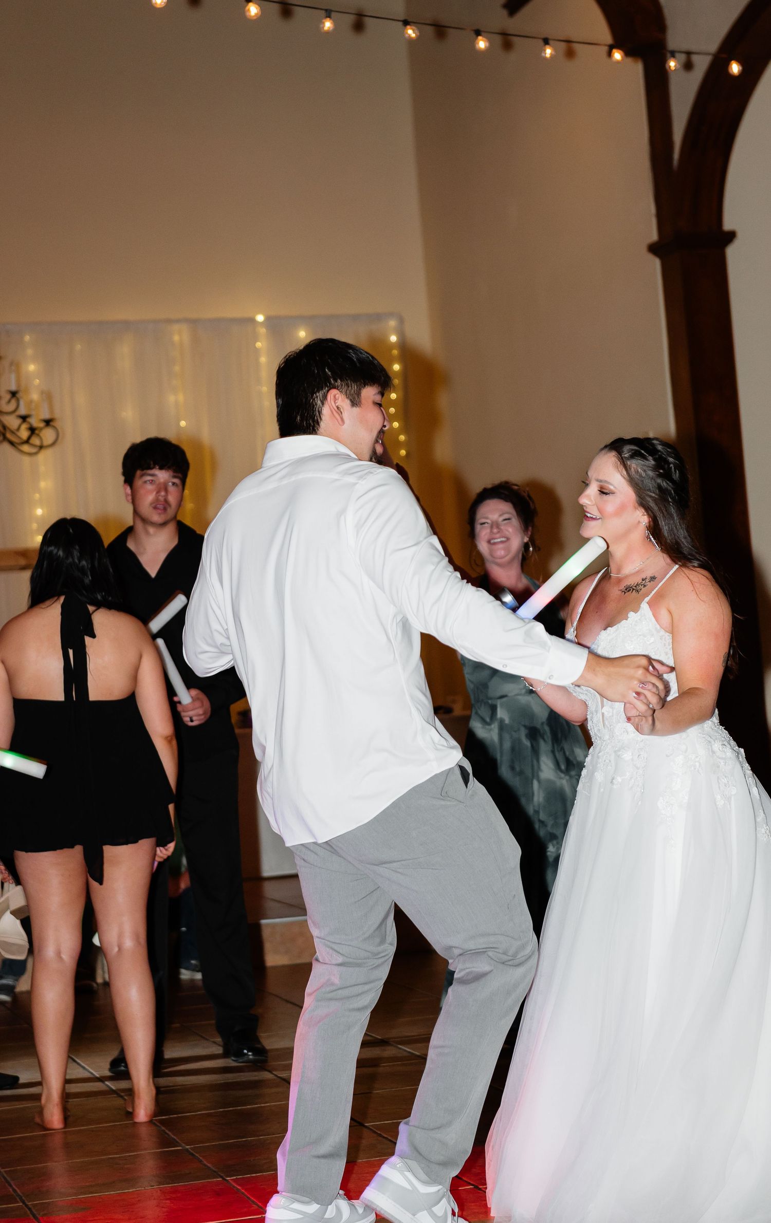 Bride and groom share a joyful first dance at their wedding reception with guests watching and string lights in background.