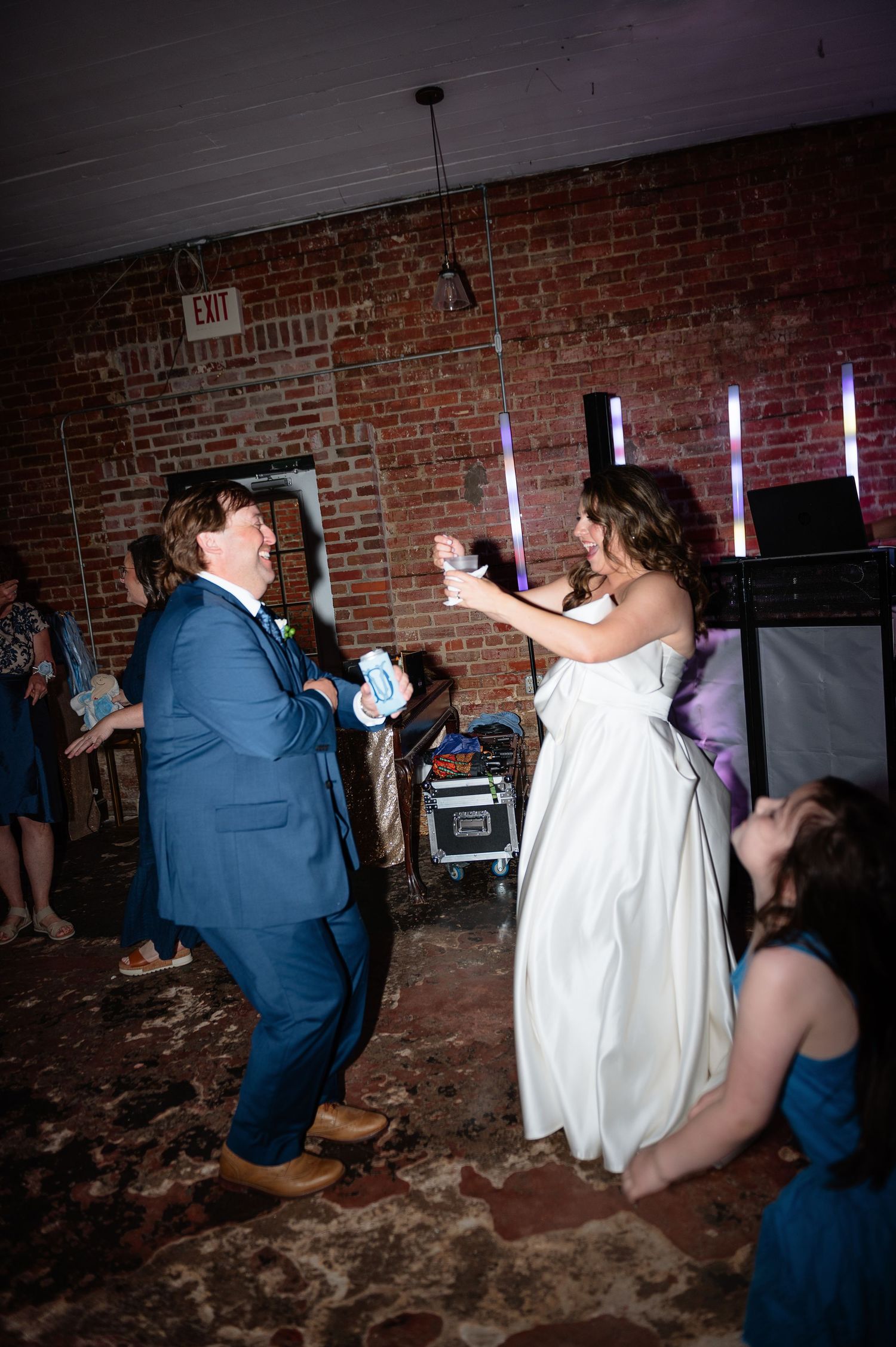 Bride and groom dancing energetically at wedding reception in rustic brick venue with colorful lights.