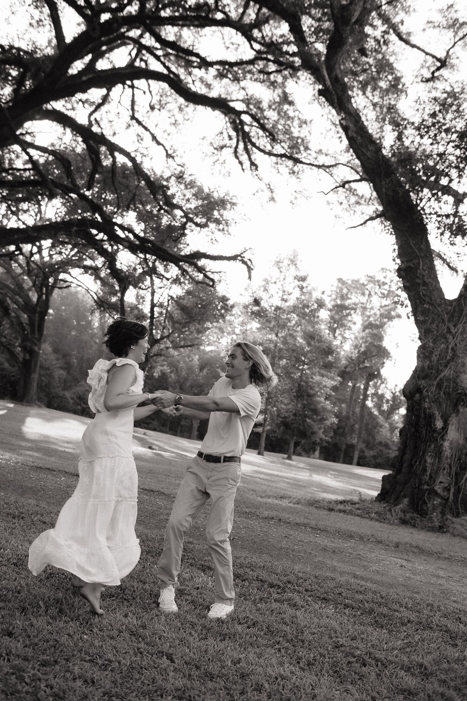 Black and white photo of a couple dancing joyfully outdoors under a large oak tree on a grassy lawn.