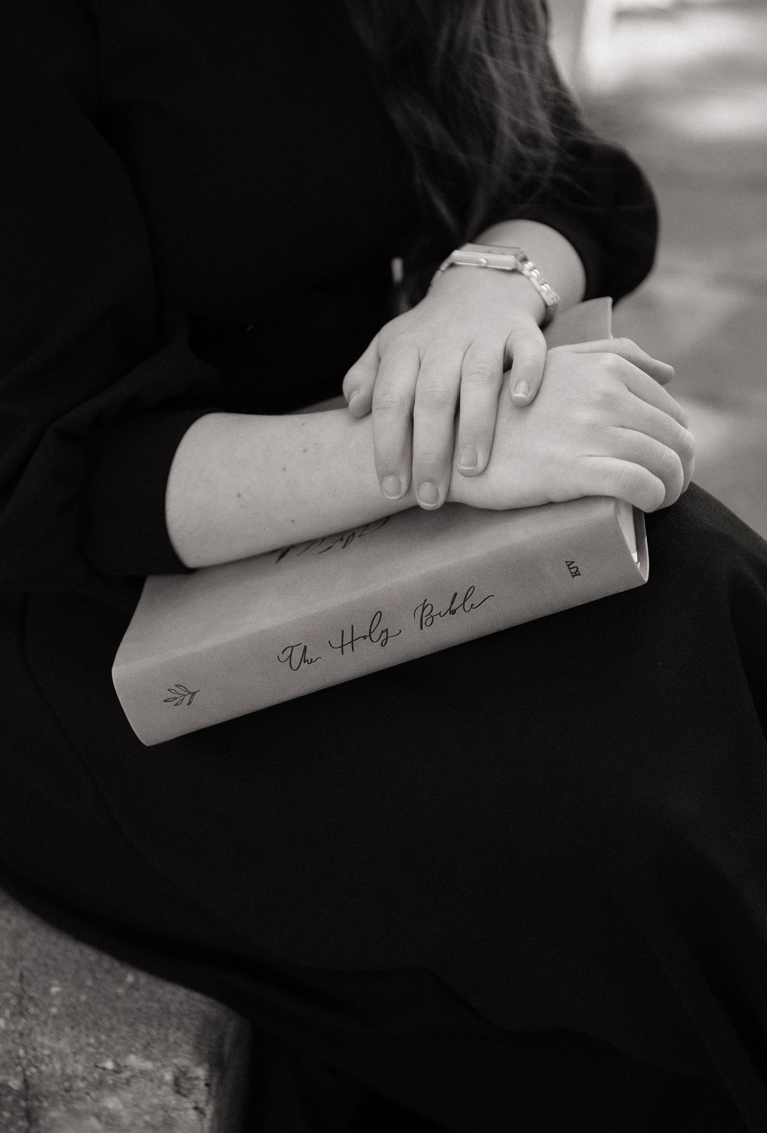 Black and white photo of a woman in black dress holding a Holy Bible on her lap with crossed hands wearing a bracelet.