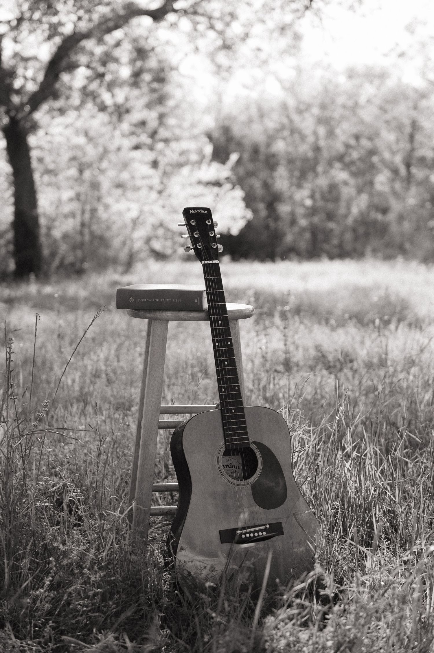 Black and white photo of an acoustic guitar leaning against a wooden stool in a grassy outdoor field.