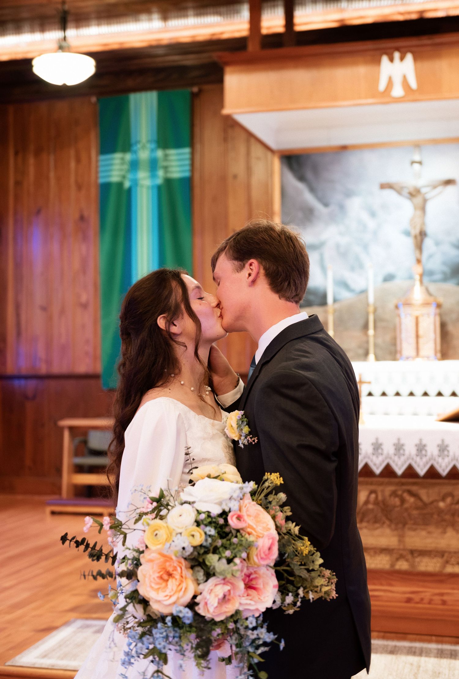 Newlyweds share first kiss at church altar, bride holding pastel floral bouquet with pink roses and blue flowers.