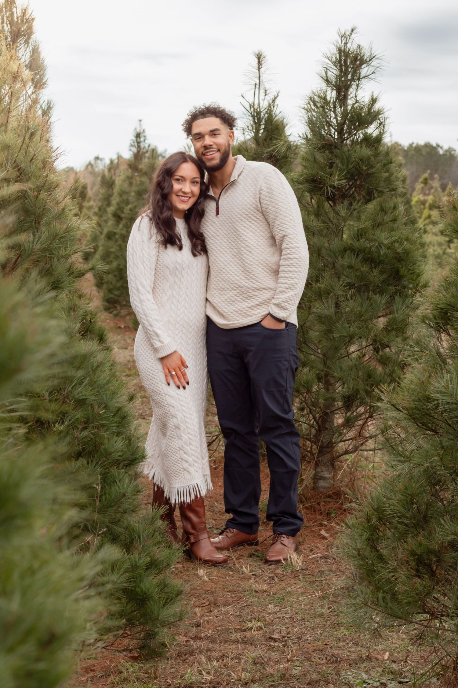 A couple in matching cream sweaters poses together among evergreen trees on a cloudy day.