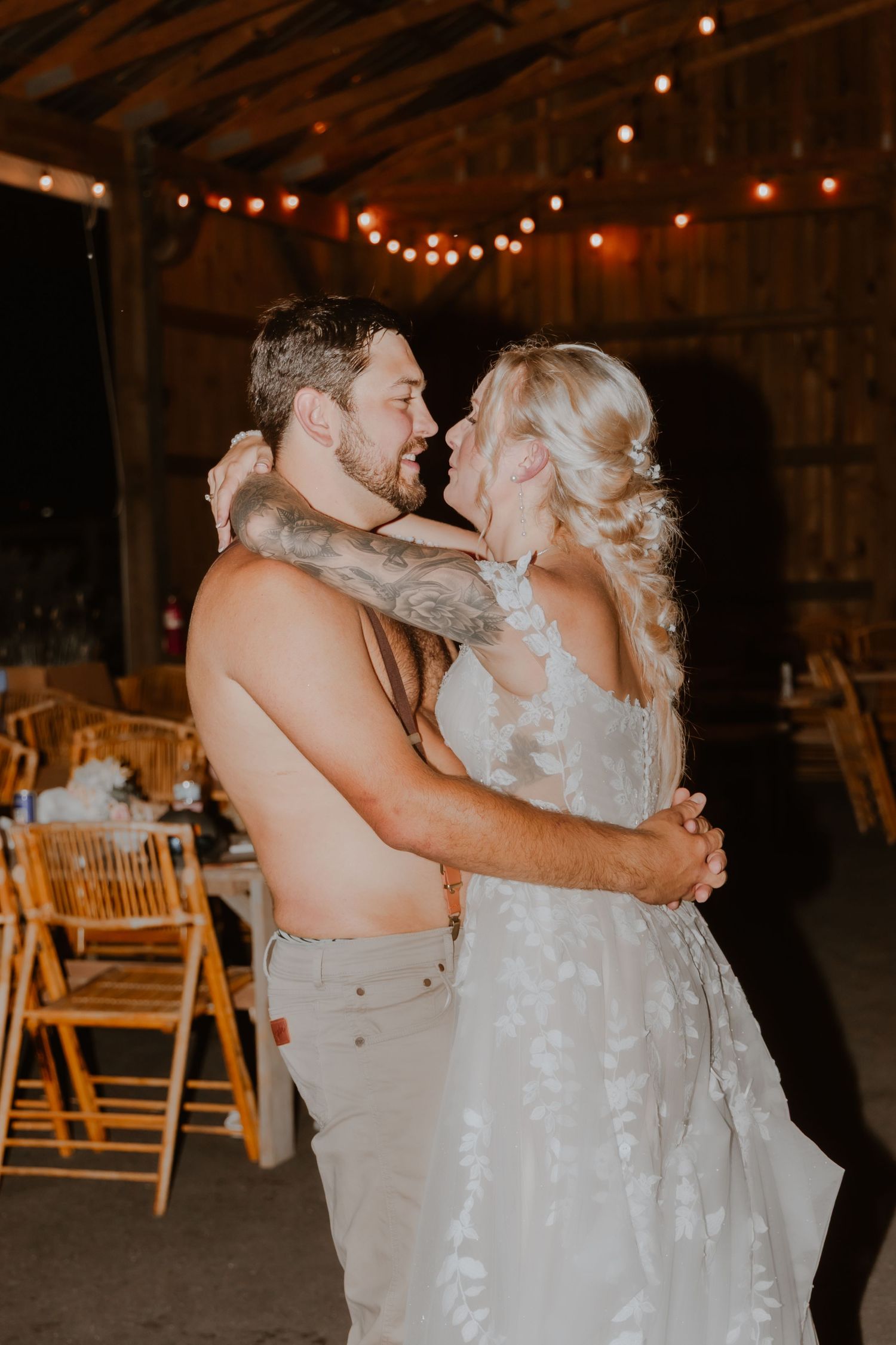 A shirtless groom and bride in a lace wedding dress share a romantic first dance in a rustic barn with string lights.