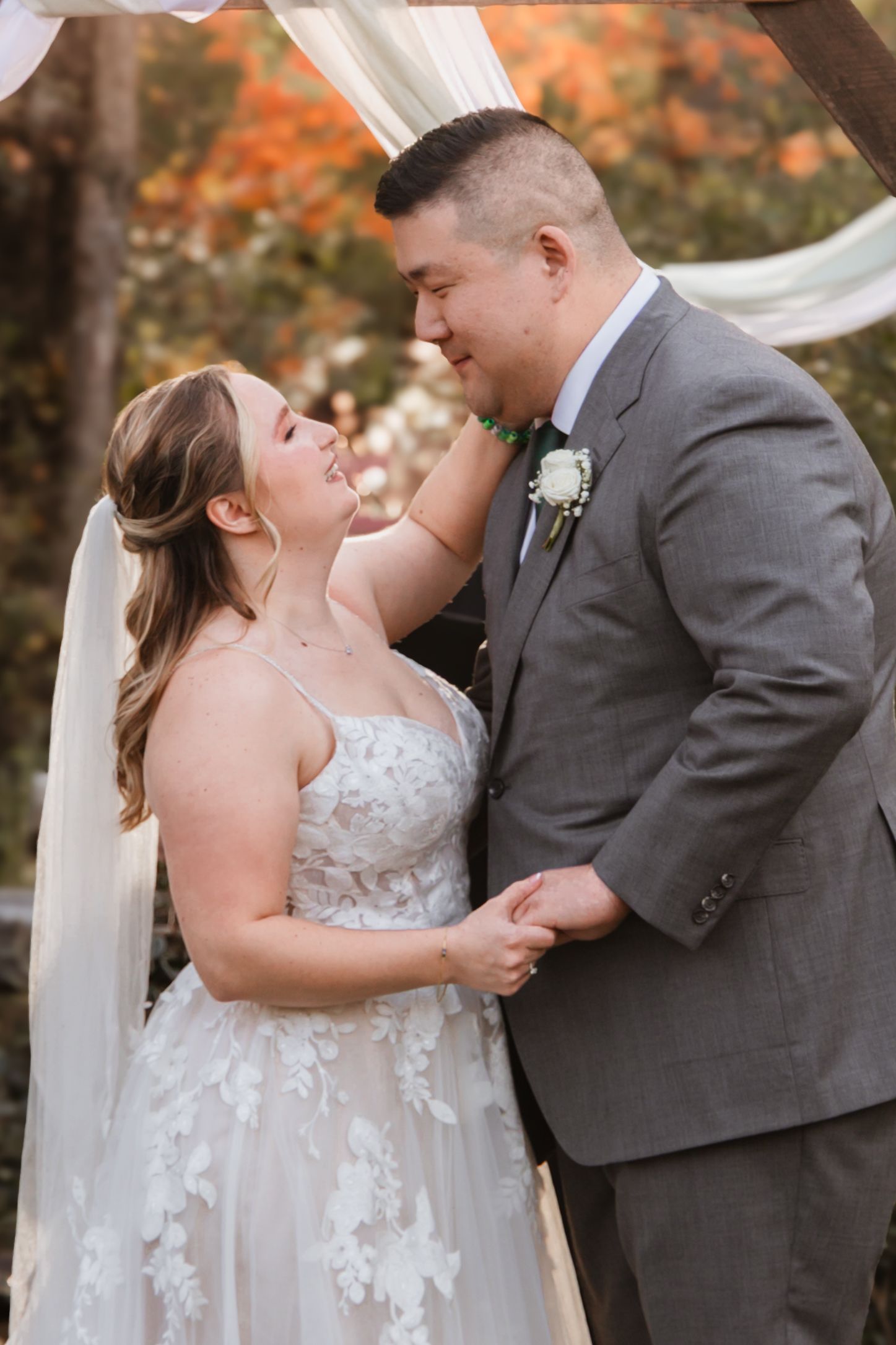 A bride and groom share a joyful moment under a floral arch with autumn foliage in the background.