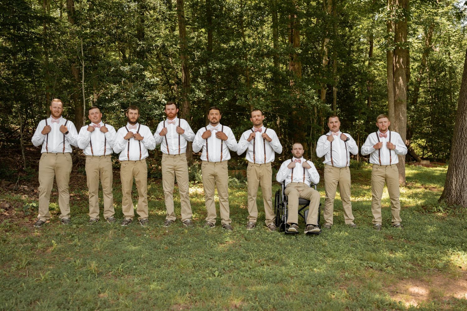 Groomsmen in khaki pants and white shirts with suspenders pose outdoors in a wooded setting for a wedding photo.
