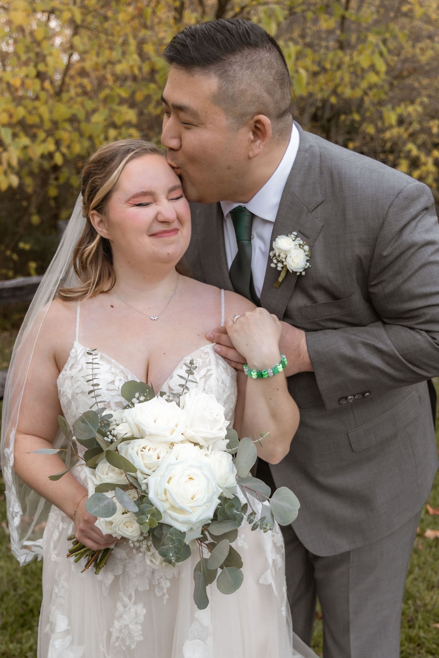 Bride in lace gown holding white bouquet with eucalyptus poses with groom in grey suit during outdoor wedding photos.