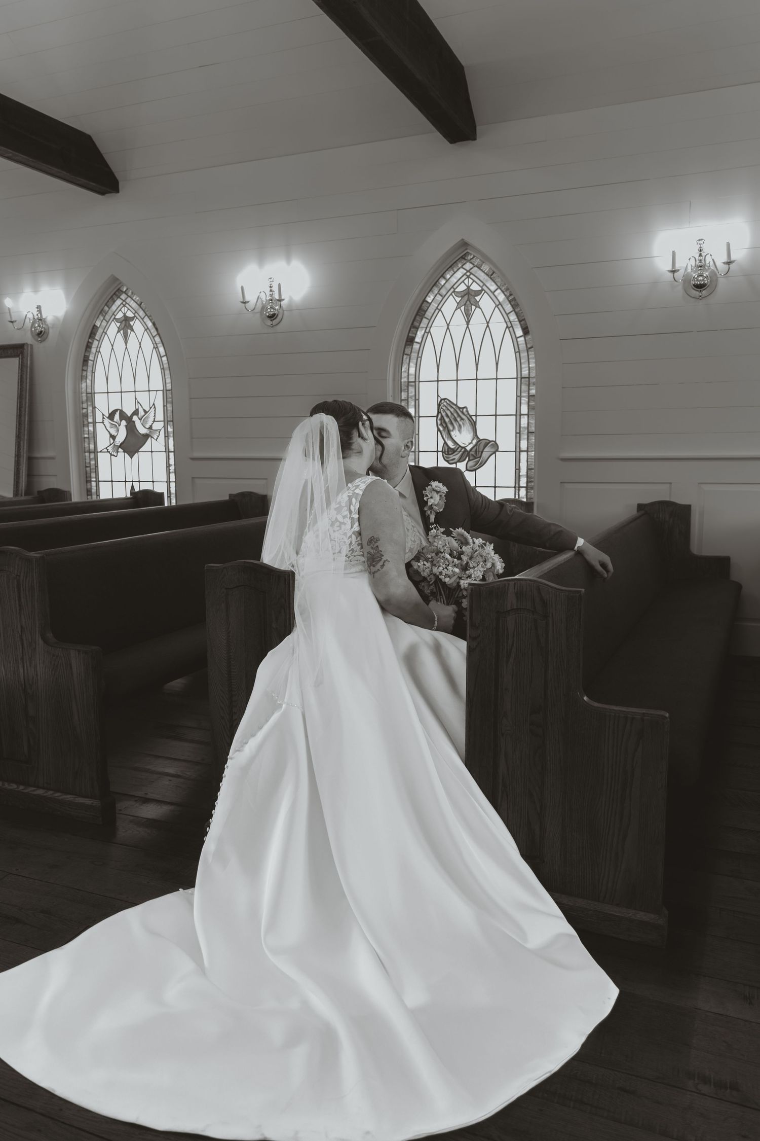 Black and white photo of a bride and groom kissing in a chapel with stained glass windows and wooden pews.