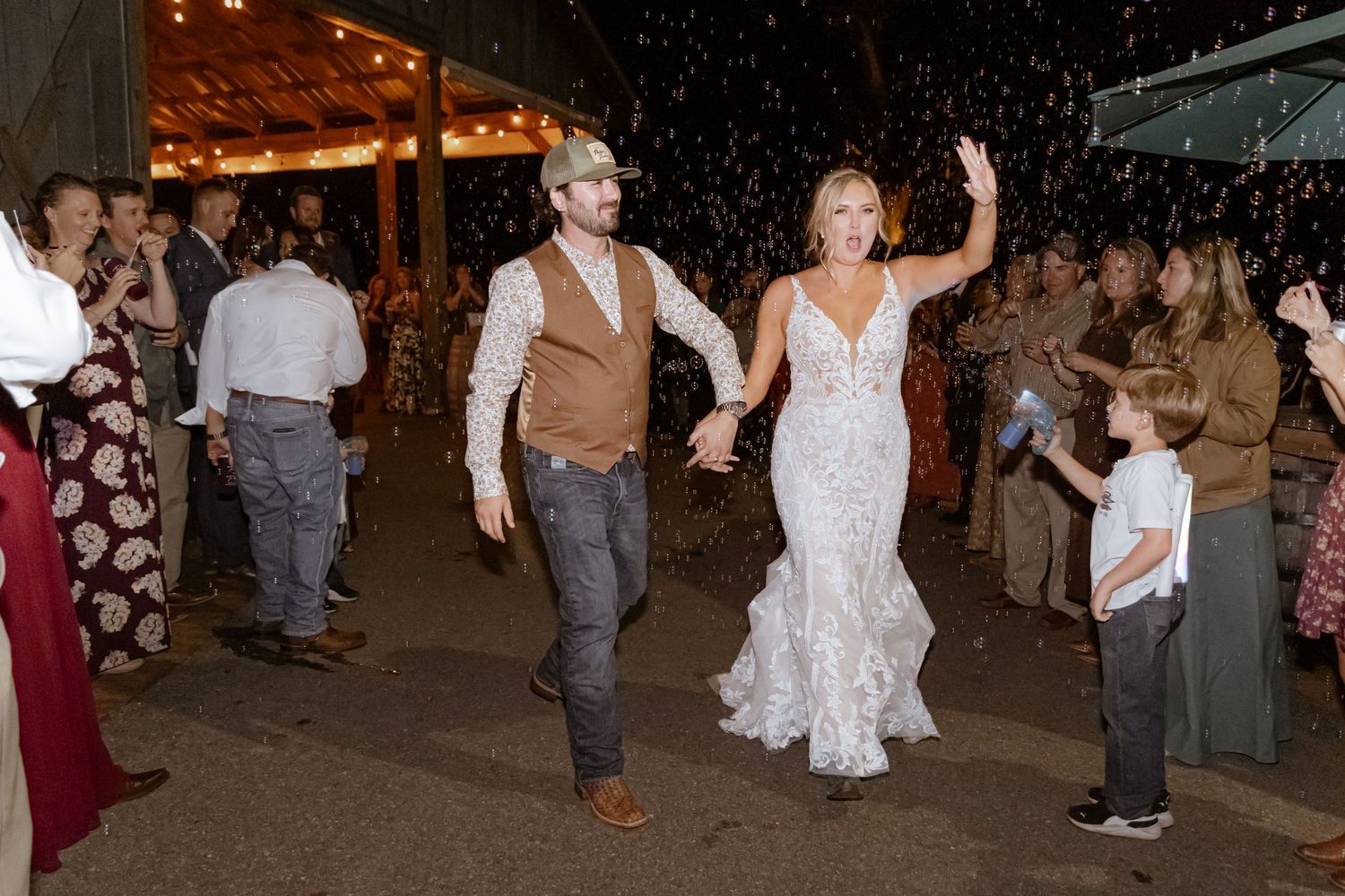Bride in lace gown and groom in brown vest exit wedding reception amid celebratory rice toss with guests cheering.