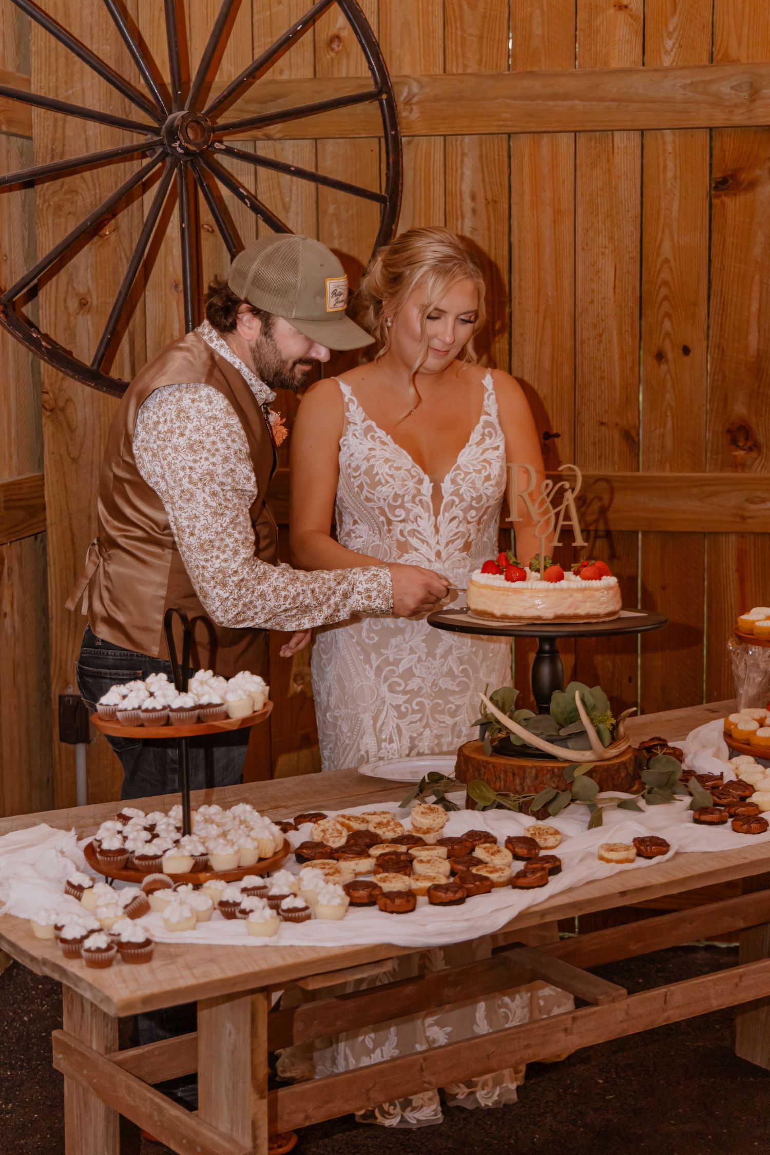 Bride and groom cut wedding cake at rustic barn reception with dessert table display.