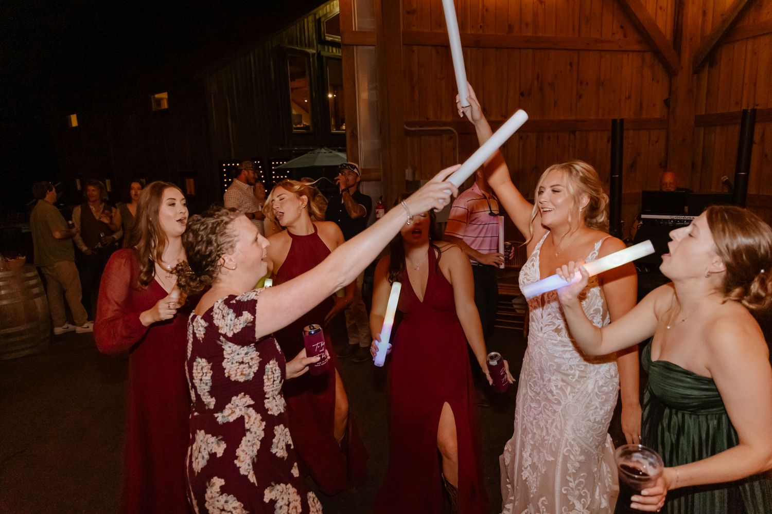 Bridesmaids in burgundy dresses celebrate at a wedding reception, waving glow sticks on the dance floor.
