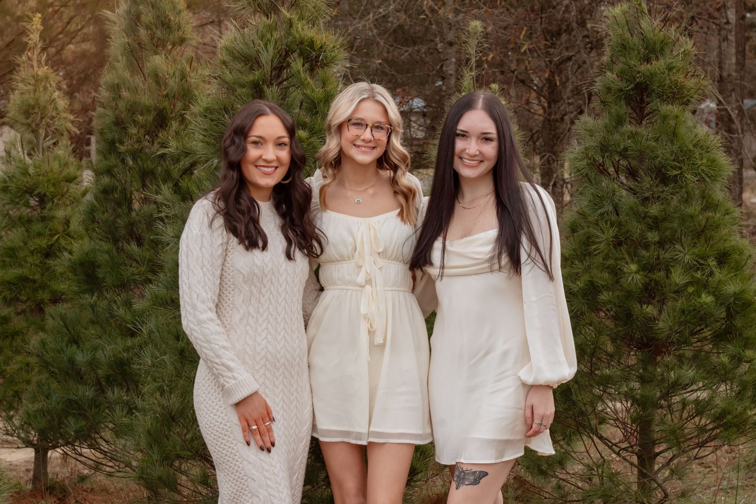 Three women in white dresses pose together at a Christmas tree farm.
