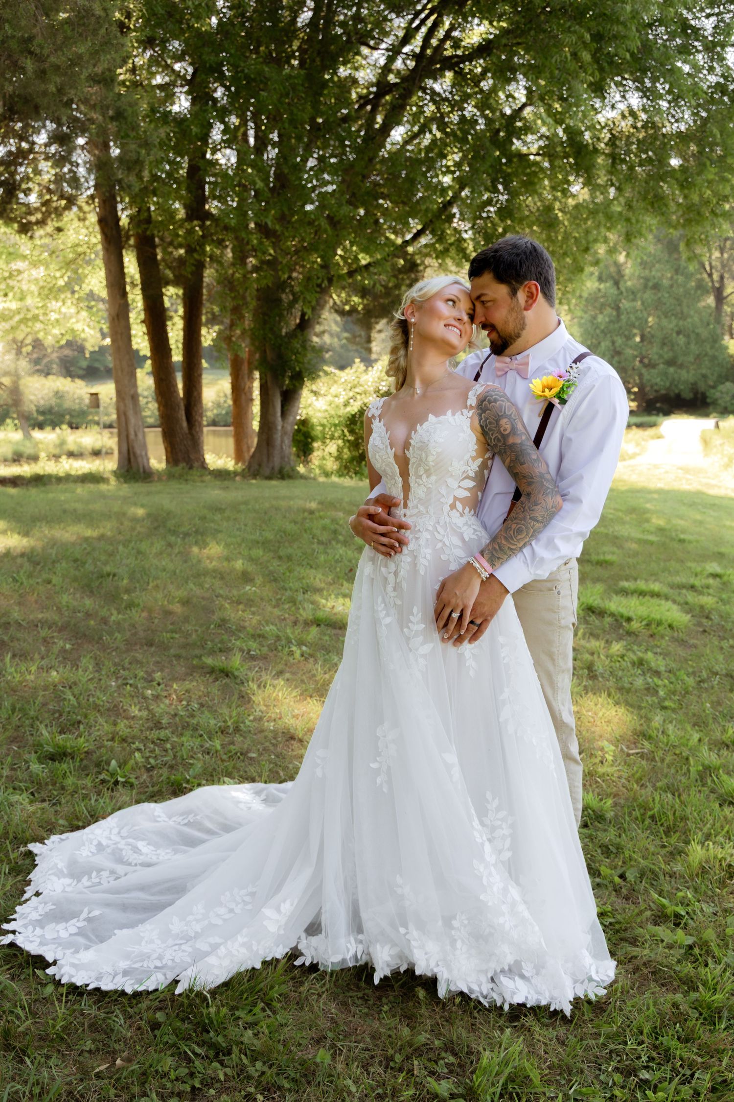 Bride in lace gown with train embraces tattooed groom among trees in sunlit garden.