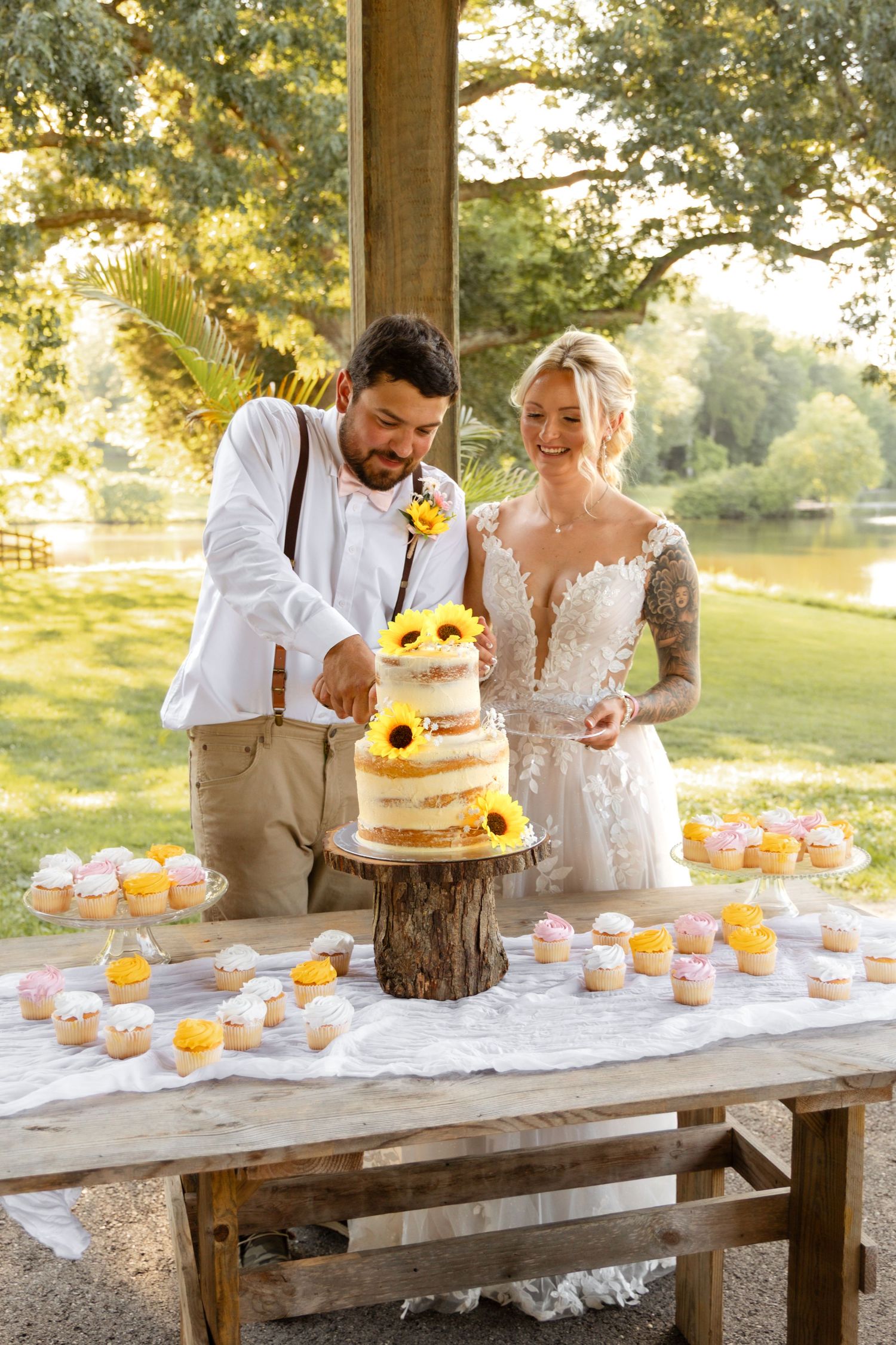 Bride and groom cut rustic naked wedding cake decorated with sunflowers at outdoor reception.