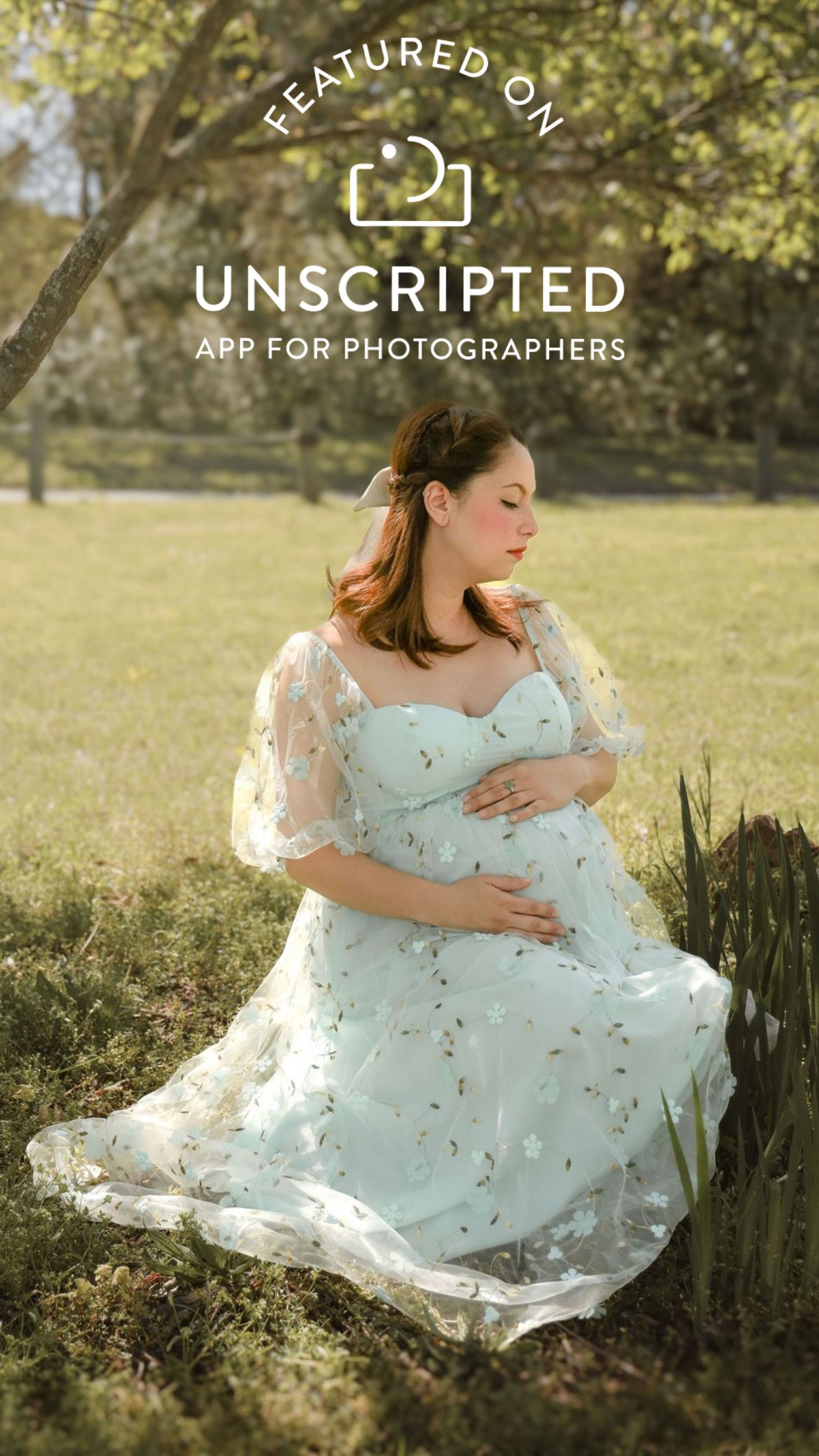 A maternity portrait of a woman in a flowing white dress sitting gracefully in a sunny meadow.
