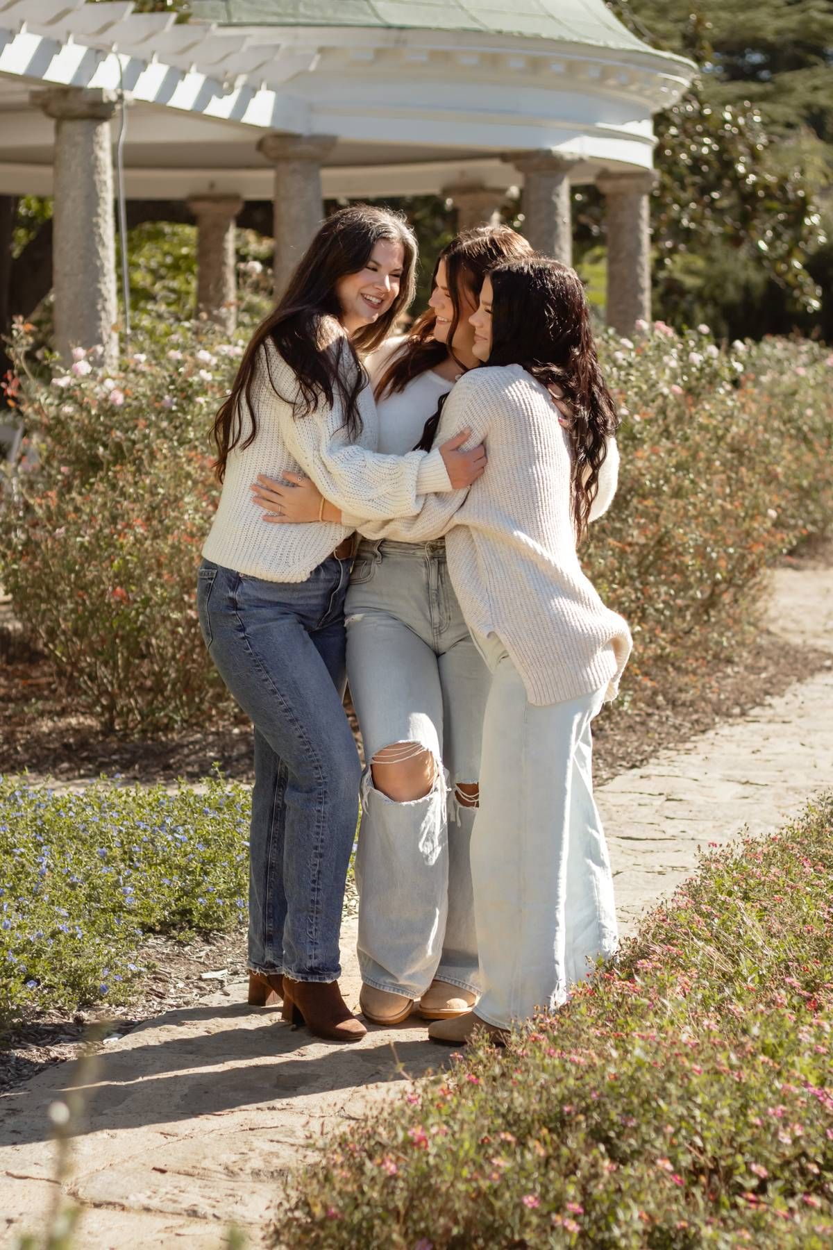 Three friends in cozy white sweaters share a warm embrace under a garden pergola on a sunny day.