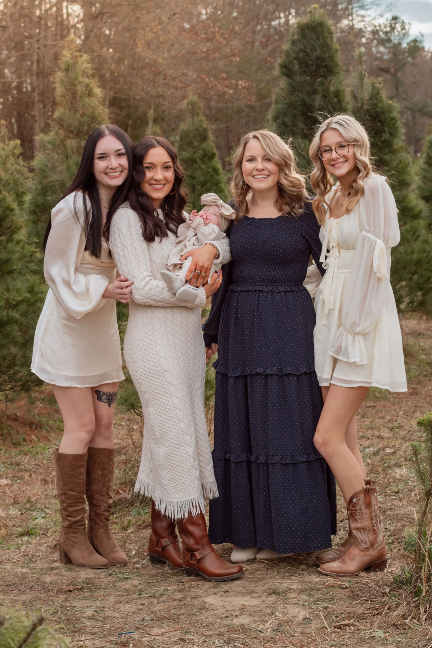 Group of women in coordinated white and navy outfits pose with a baby at a tree farm.