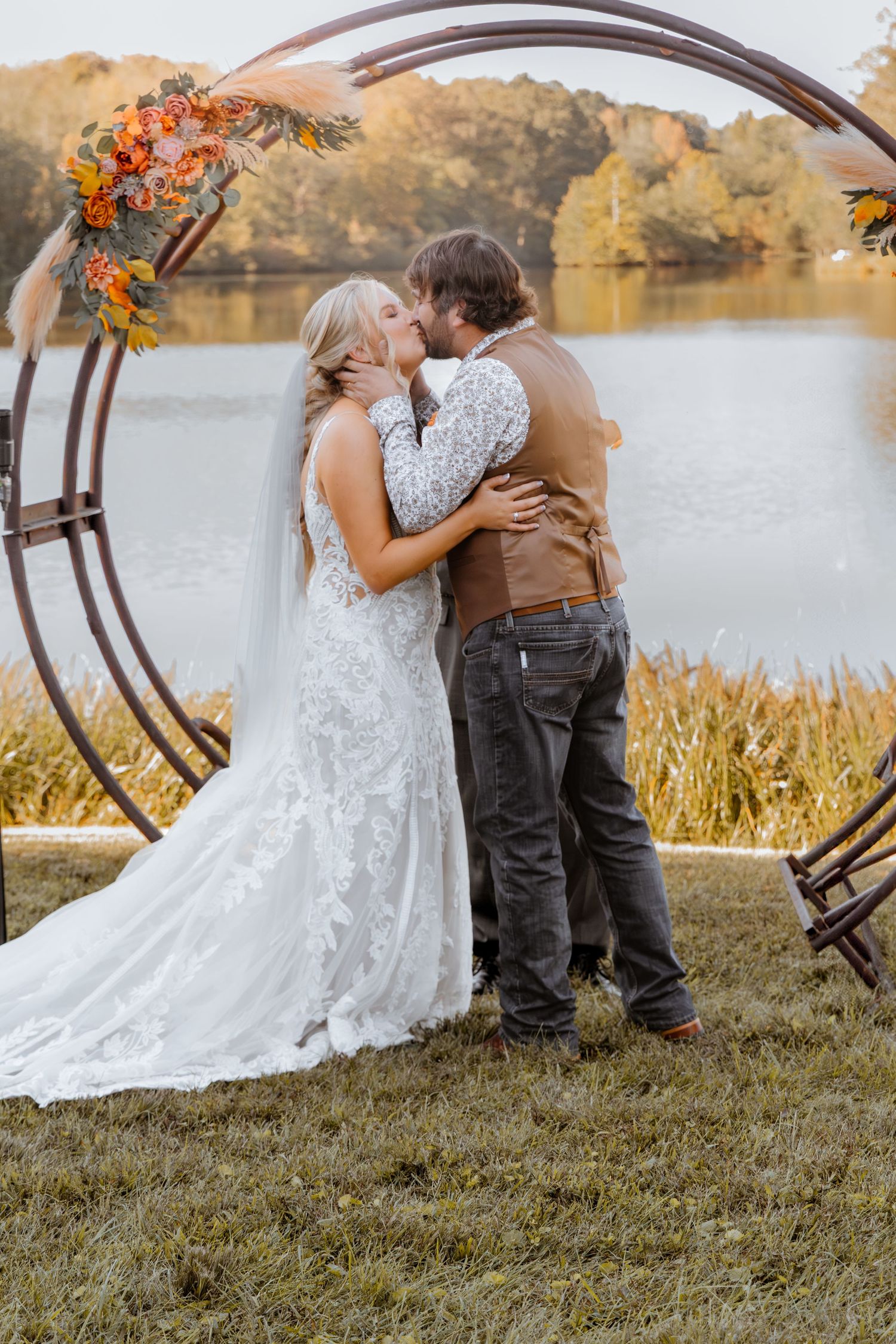 Wedding couple sharing an intimate moment under a wooden arch by a lakeside during autumn.