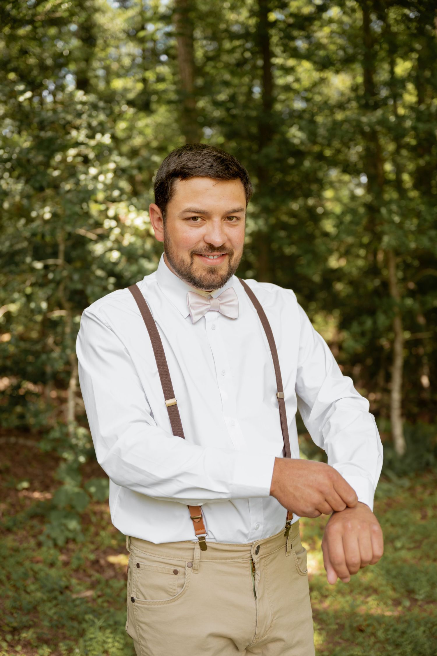 Smiling groom in white shirt, brown suspenders, and bow tie adjusting cufflinks outdoors in a forest setting.