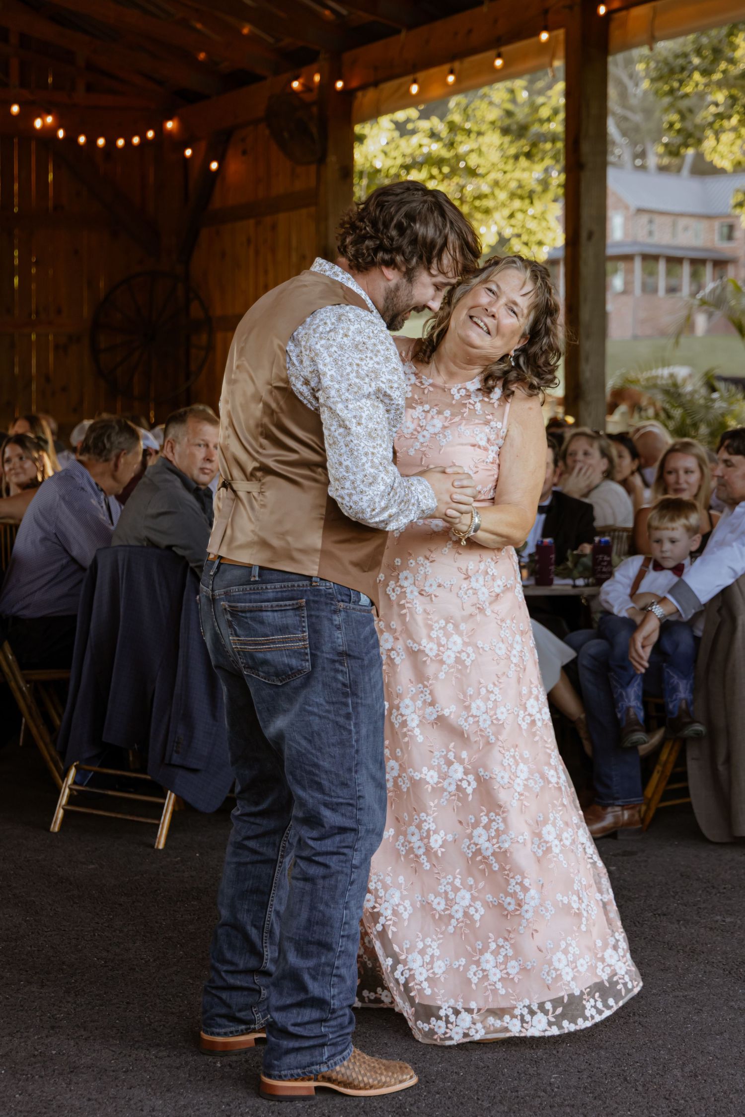 Groom shares joyful mother-son dance at rustic barn wedding reception with guests seated.