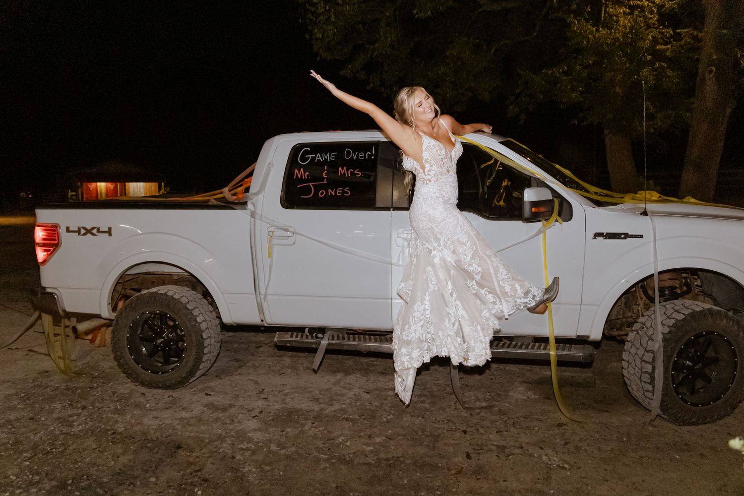 Bride in lace wedding dress posing on white Ford F-150 truck at night with yellow straps.