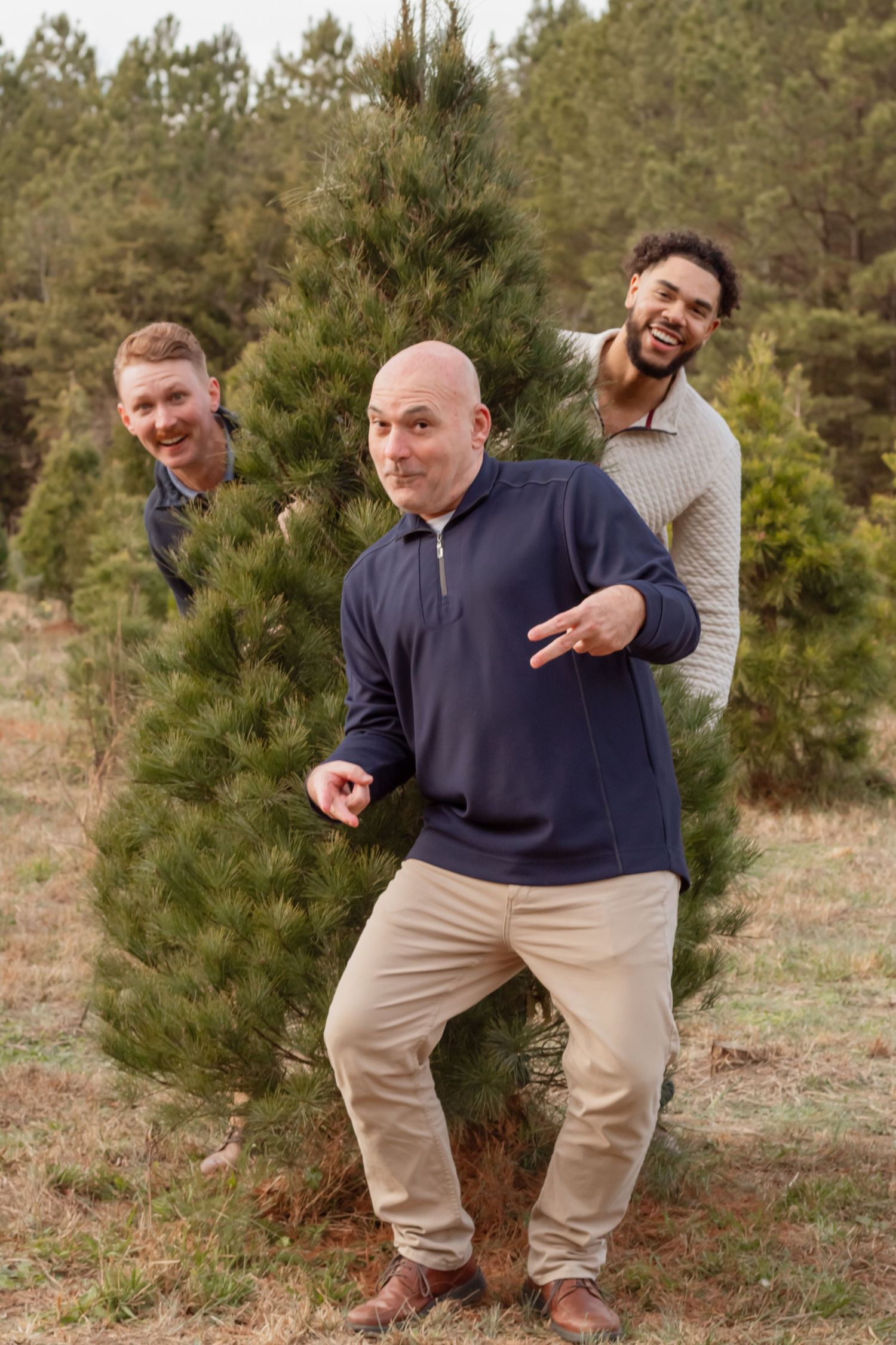 Three men pose playfully between evergreen trees during a photo session.