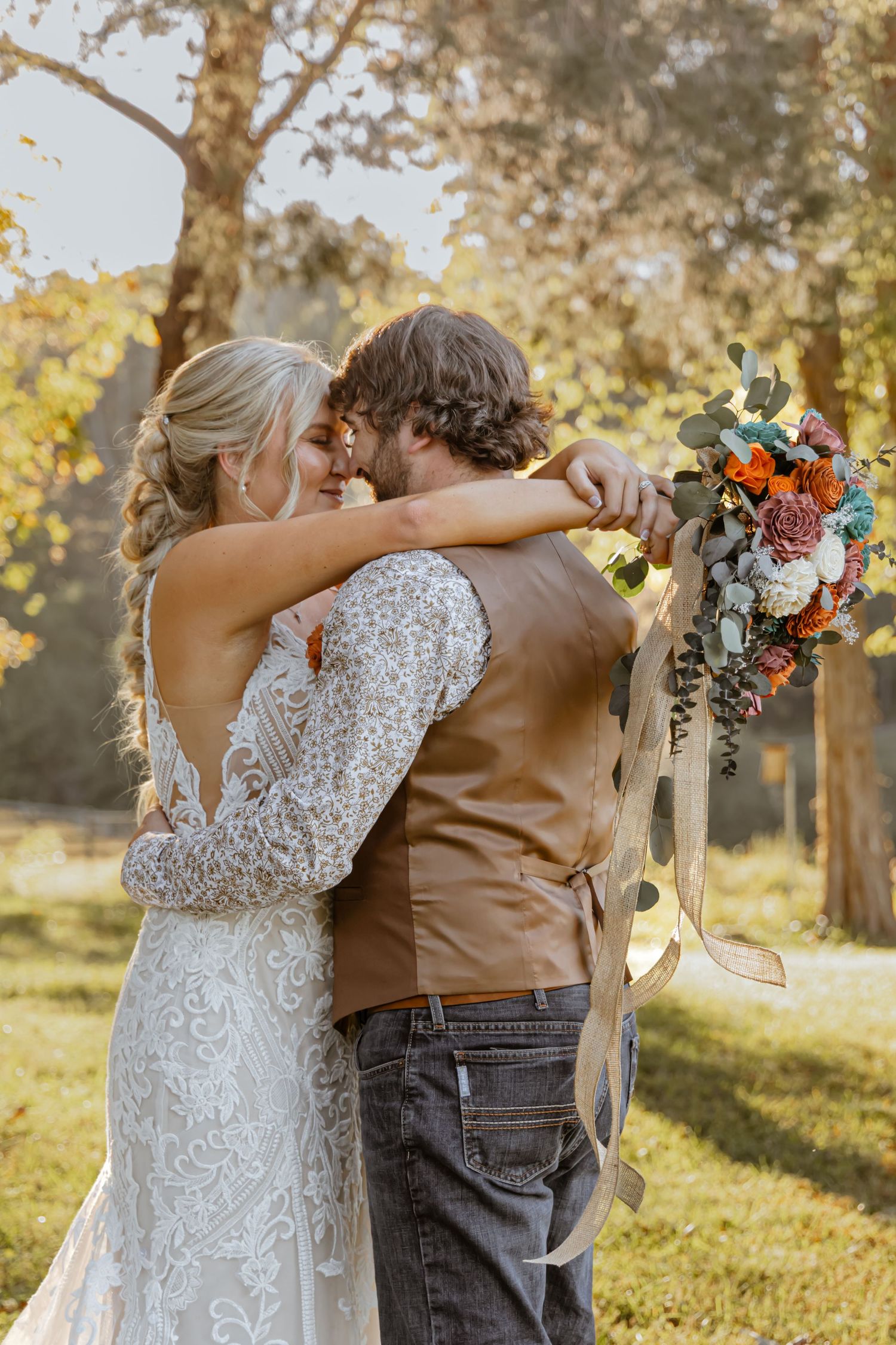 Bride and groom embrace outdoors in autumn, she holds colorful bouquet in lace gown, he wears brown vest and jeans.