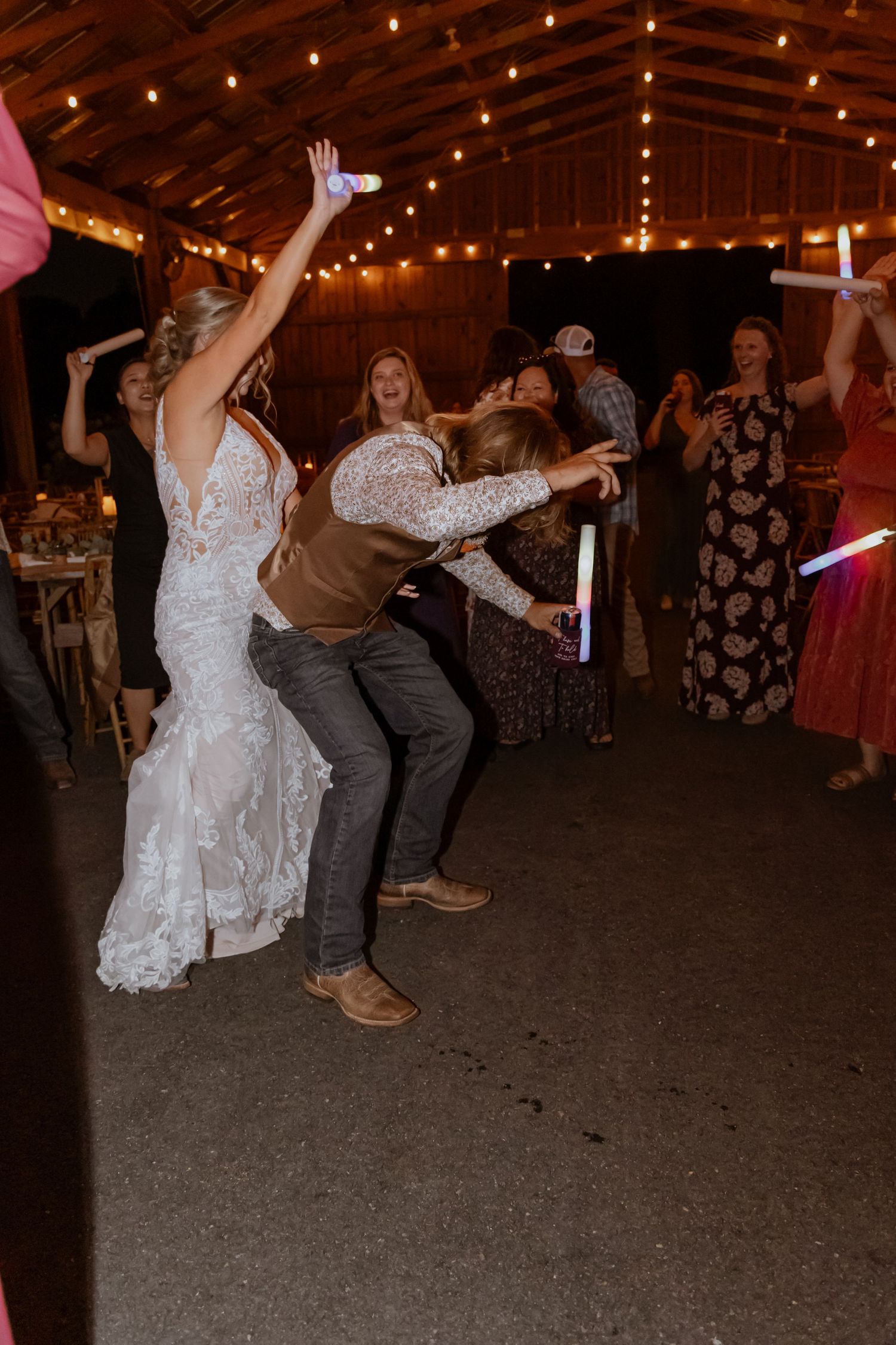 Bride in lace gown dancing with guests at rustic barn wedding reception with string lights and glow sticks.