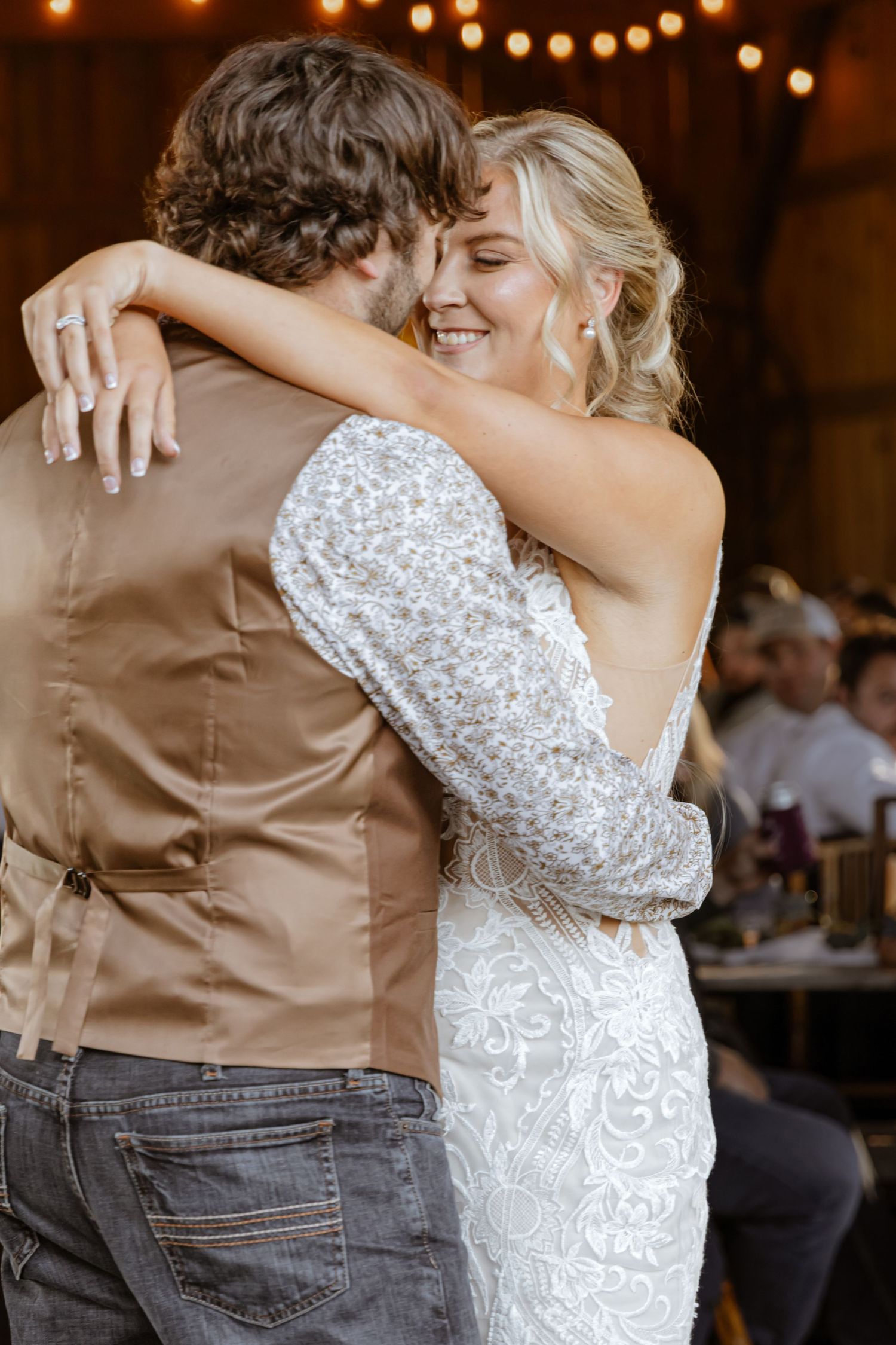 Bride in lace gown smiles joyfully while sharing first dance with groom in tan vest at rustic barn wedding.