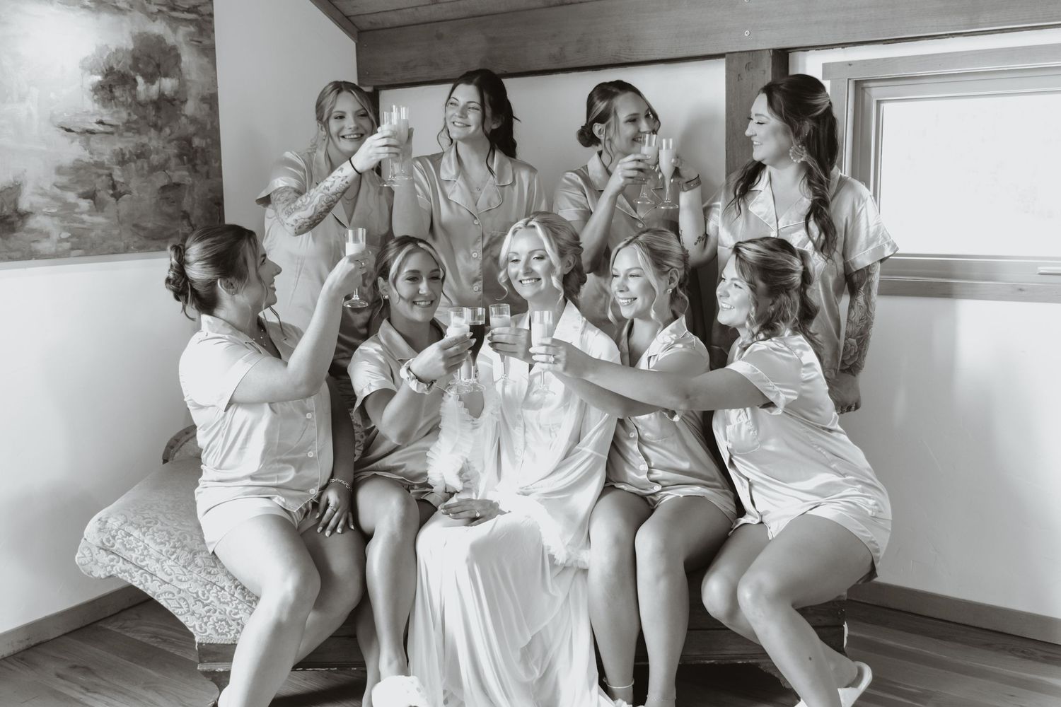 Bride and bridesmaids in robes toasting with wine glasses in a black and white wedding preparation photo.