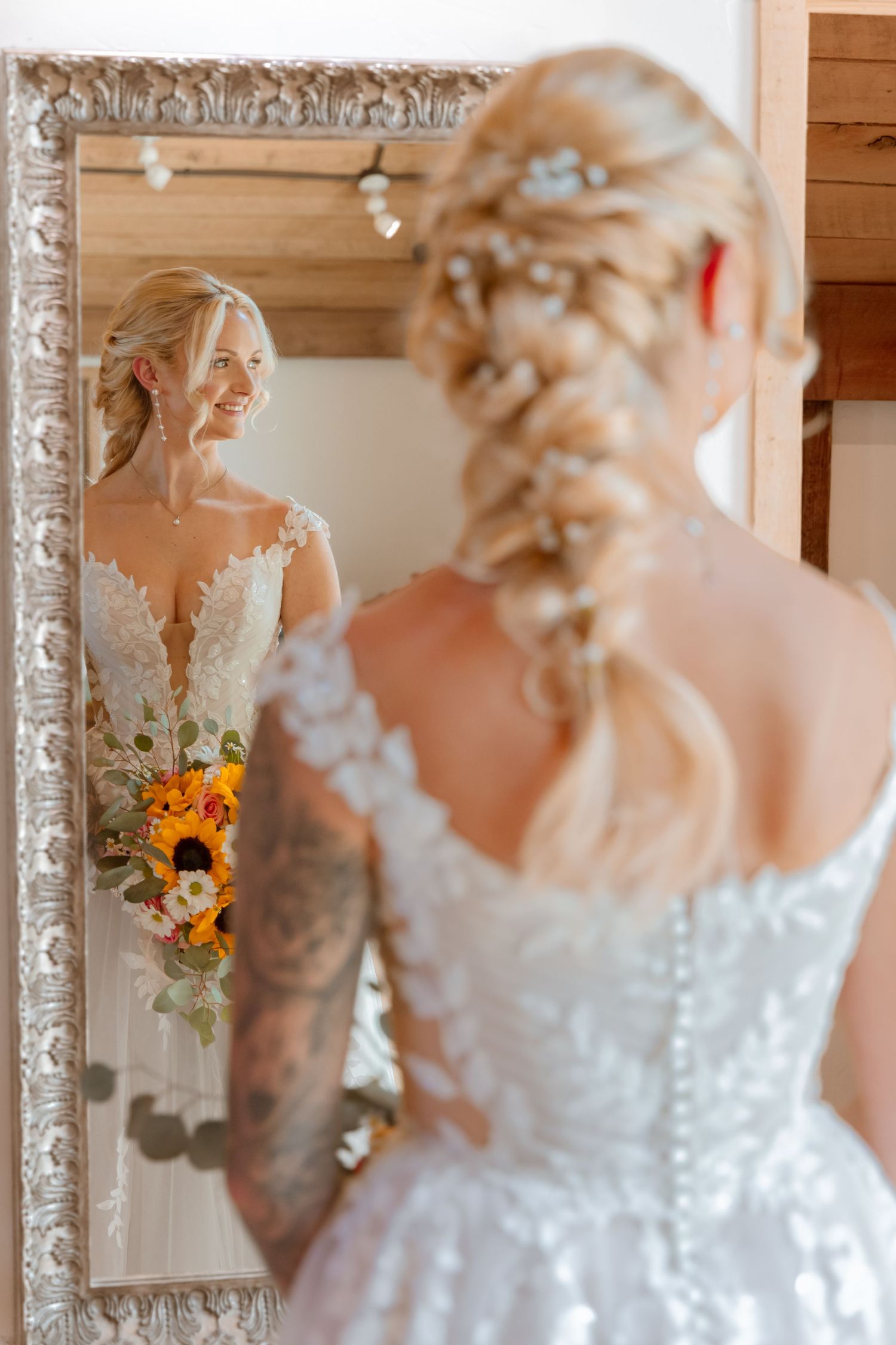 Smiling bride in lace wedding dress with braided updo holds sunflower bouquet, reflected in ornate silver mirror.