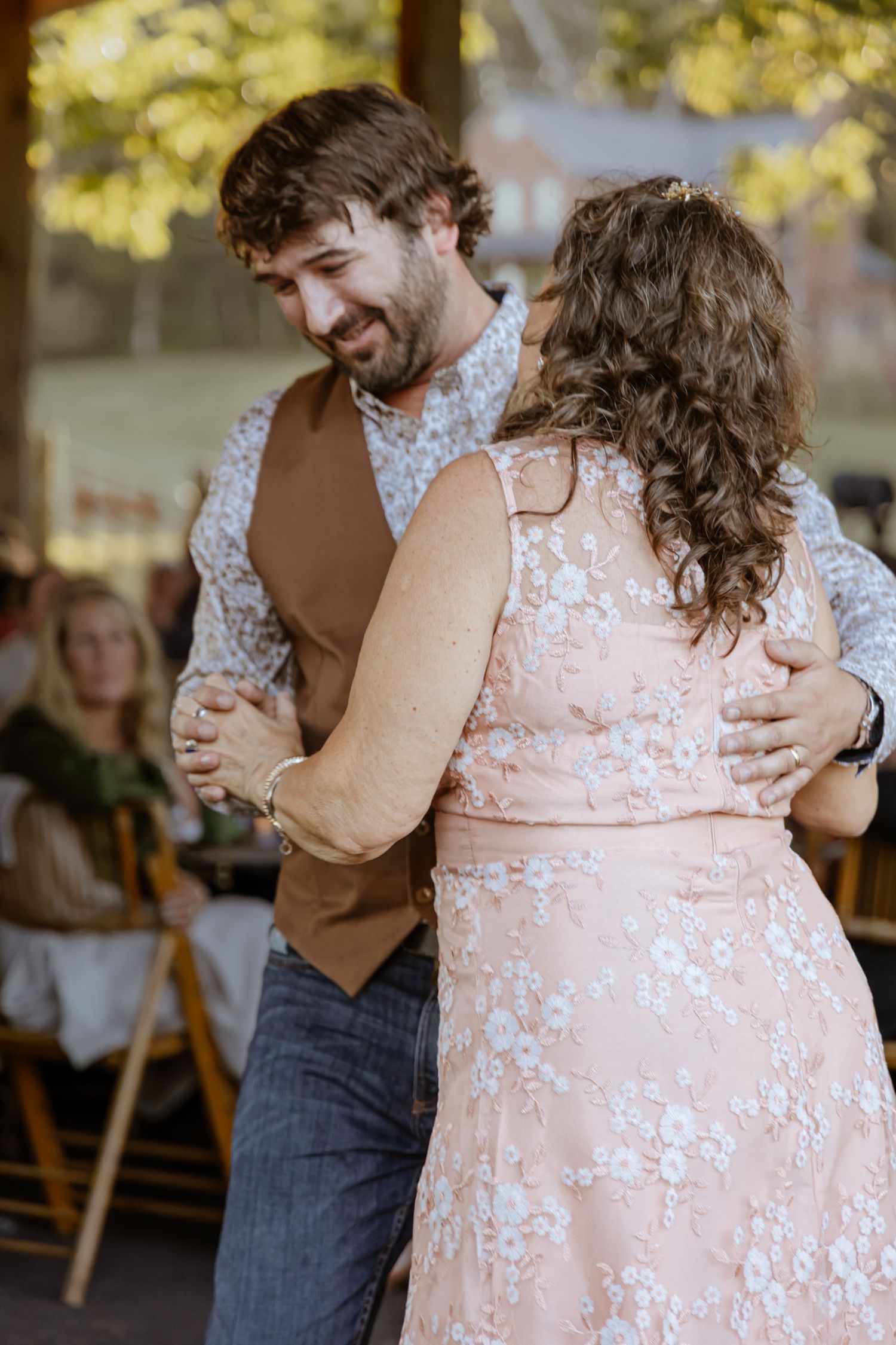 A smiling groom in a brown vest dances with a woman in a pink floral dress at an outdoor wedding reception.