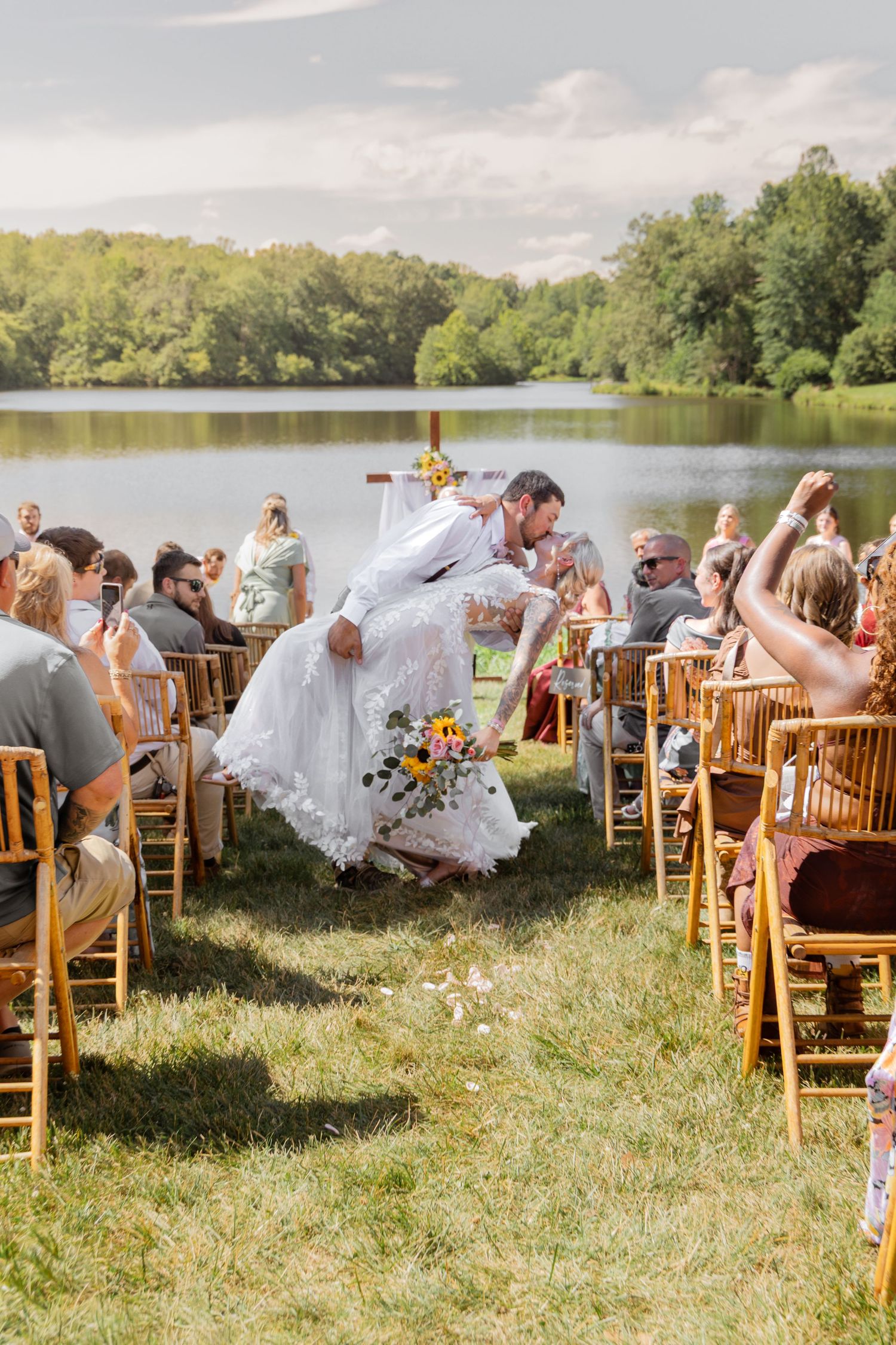 Groom dips bride during outdoor wedding ceremony by a lake, guests cheering, lush greenery backdrop.
