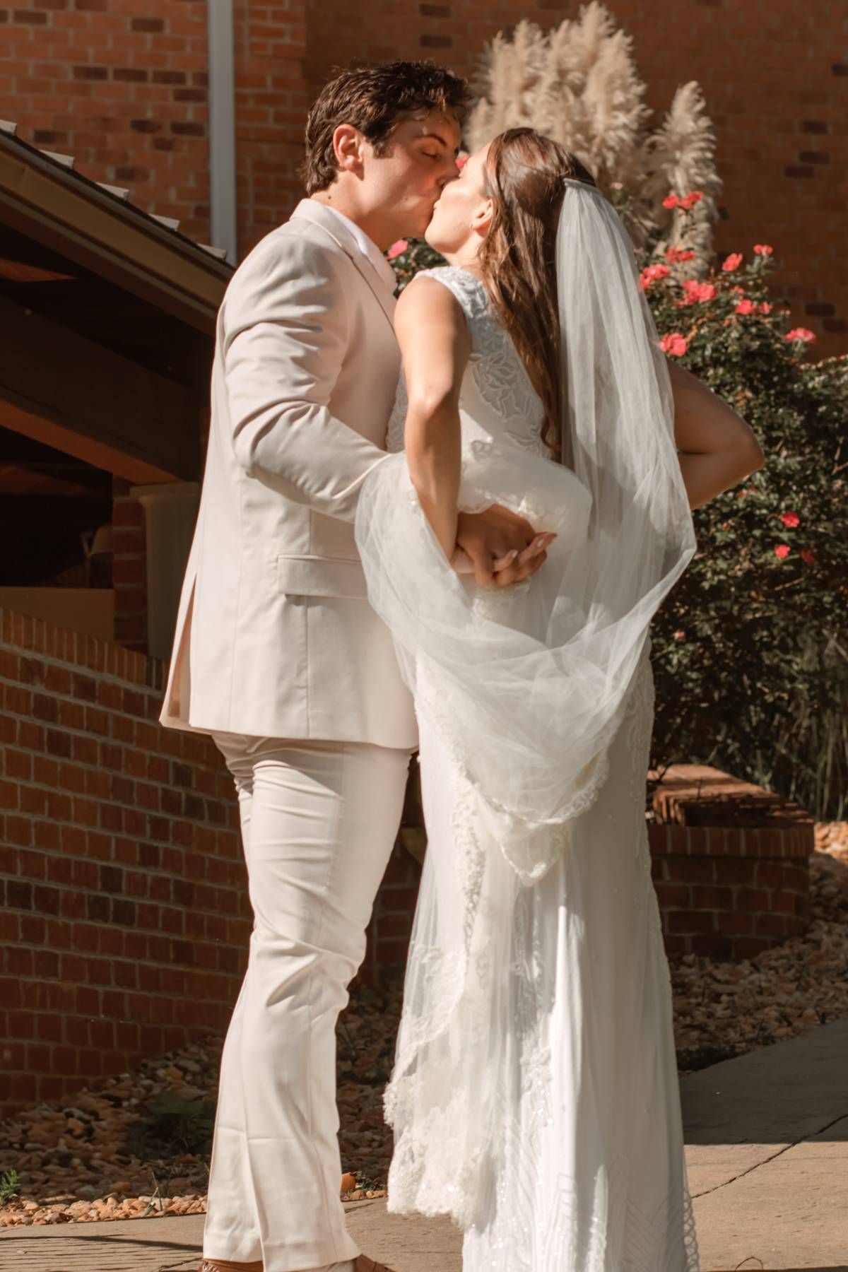 A couple in white attire shares romantic moments against a brick building with blooming roses.