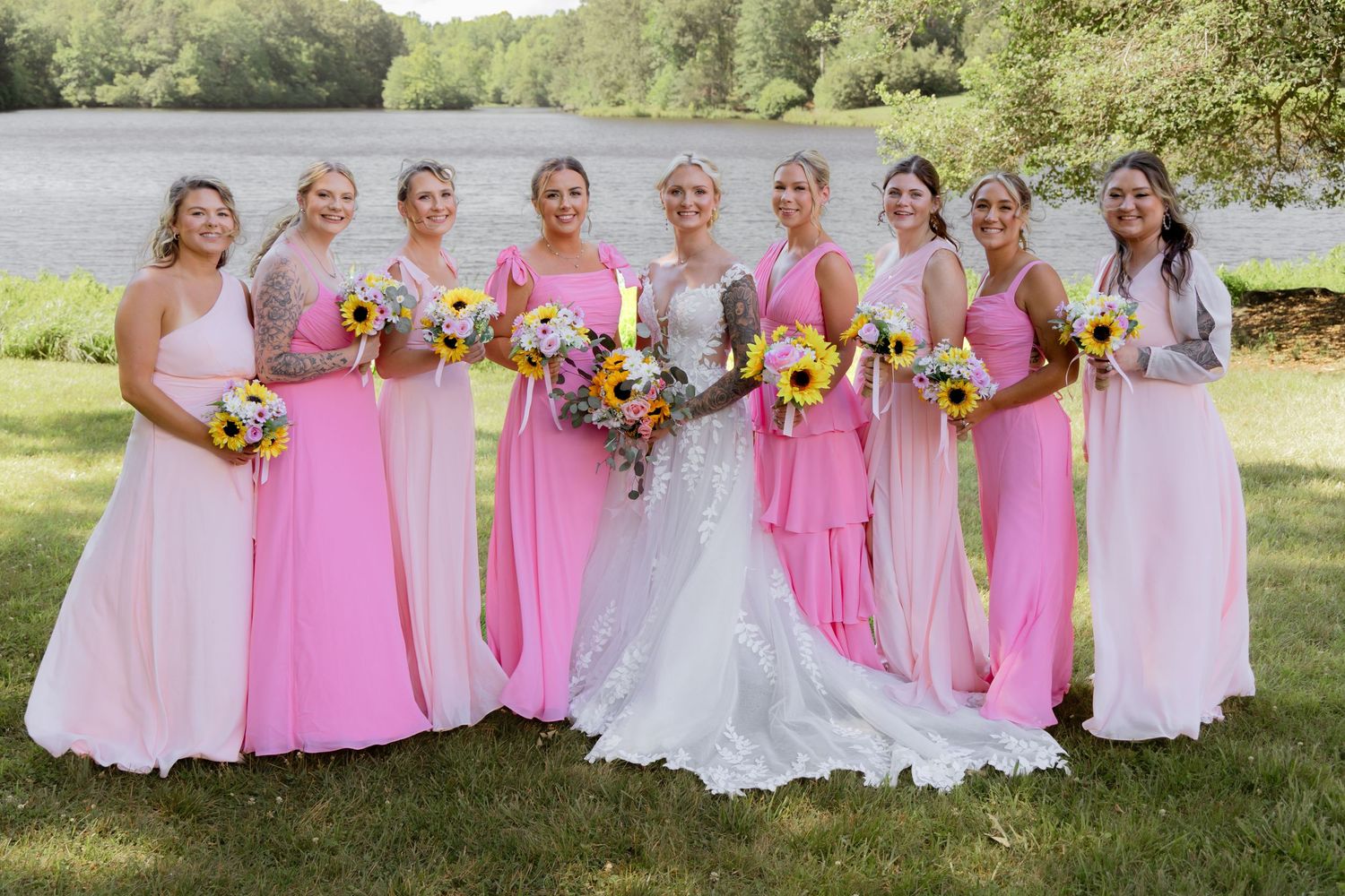 Bride in white gown poses with eight bridesmaids in pink dresses holding sunflower bouquets outdoors near a lake.