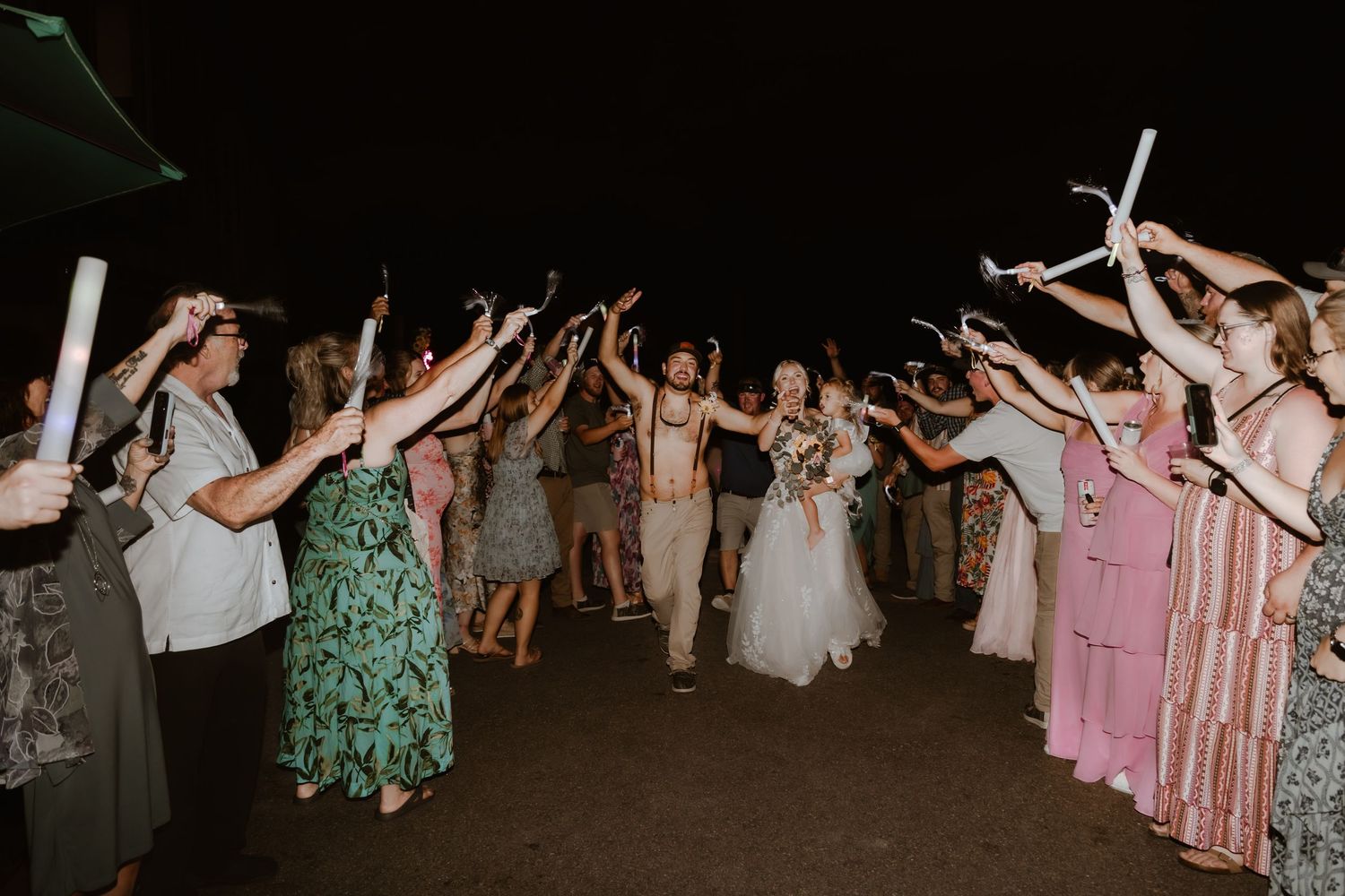 Newlyweds walk through sparkler exit tunnel surrounded by cheering wedding guests at night celebration.