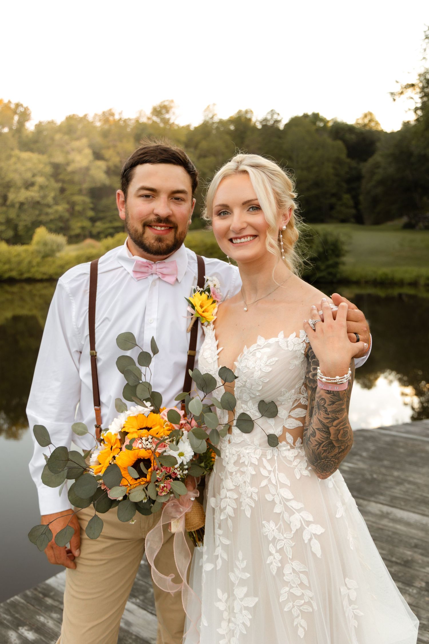 Smiling bride and groom pose on wooden dock, she displays ring while holding sunflower bouquet at sunset.