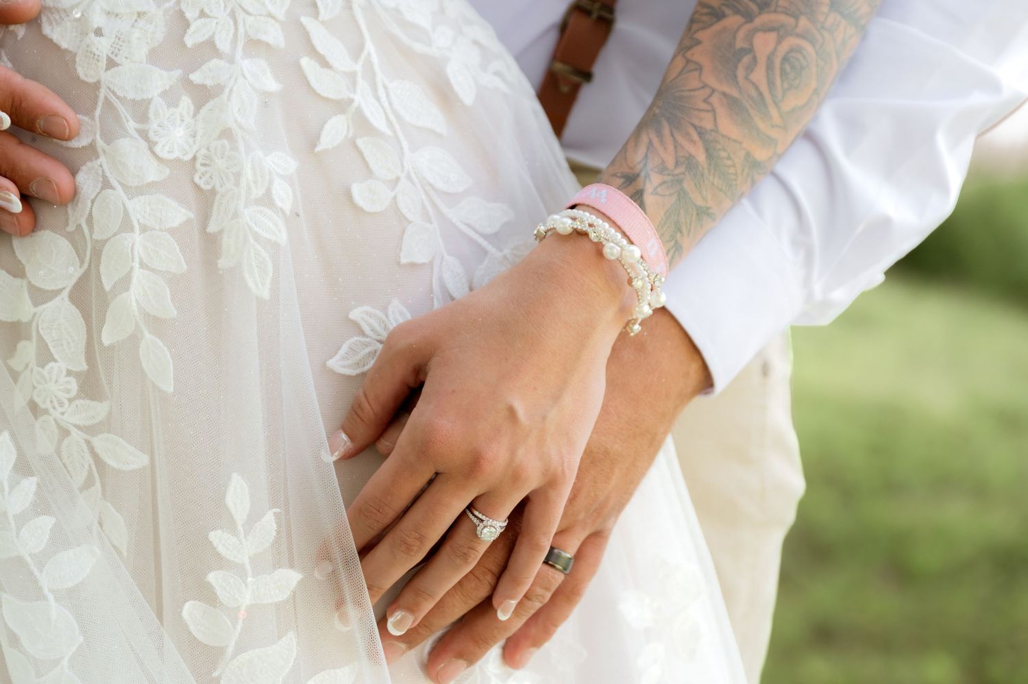 Close-up of bride and groom hands showing wedding rings, with tattooed groom arm around bride in lace dress.